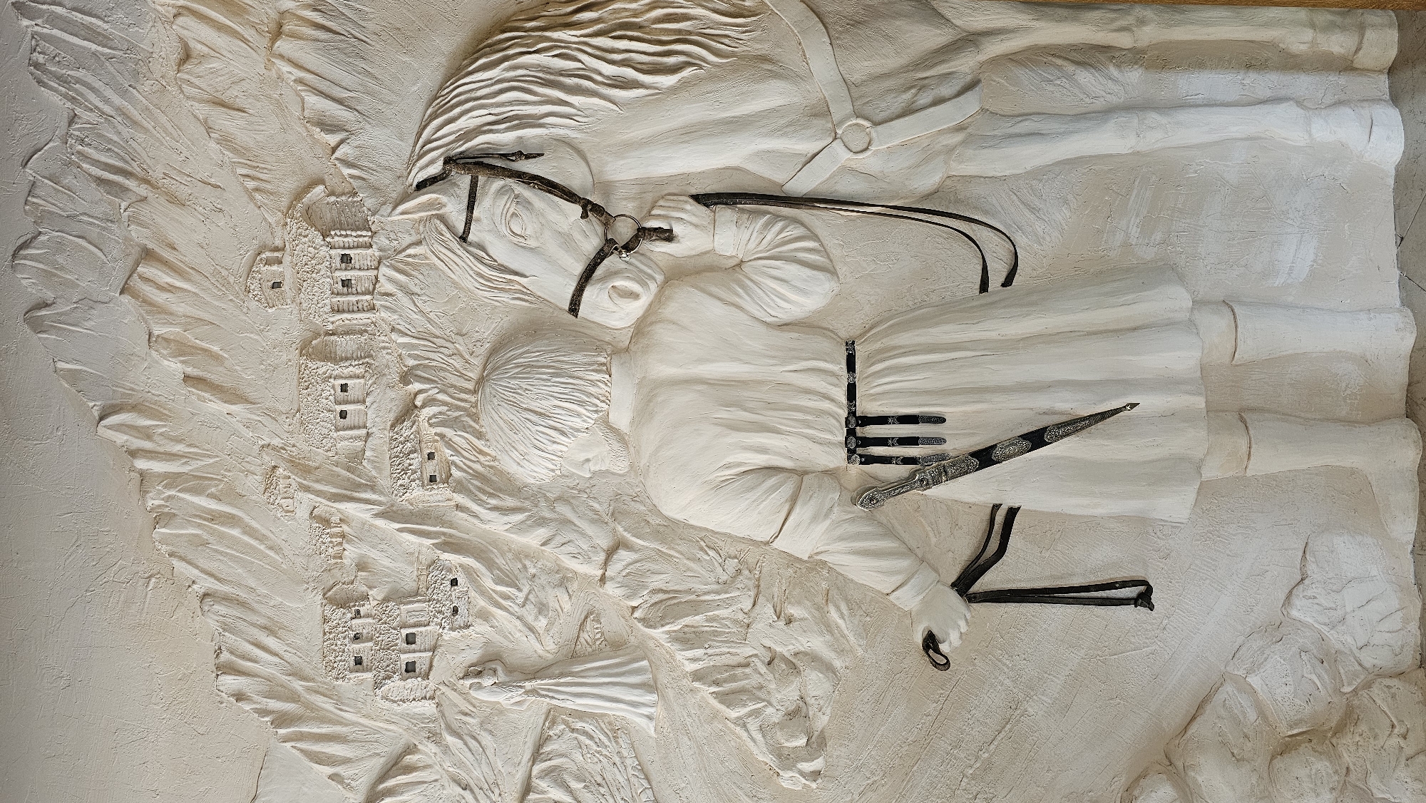 This is a relief sculpture made of light-colored material, depicting a scene set in a mountainous, village-like environment. In the foreground, a person stands facing away from the viewer, holding the reins of a white horse. The person is dressed in traditional attire, including a long robe, a patterned belt with what appears to be a dagger tucked into it, and high boots. The horse is saddled and bridled, its head turned slightly towards the person.
In the background, the mountainous terrain is textured to resemble rough rock faces and steep slopes. Clustered on these slopes are small, rustic buildings with thatched roofs, suggesting a remote village or settlement. To the far left of the frame, a solitary figure in a long, flowing dress stands on a path amidst the rocky landscape. The overall impression is a historical or folkloric scene, evoking a sense of tradition and a connection to nature. The location in the city of Красный Курган, Russia, is not depicted in the artwork itself, which presents a generic mountainous village scene.