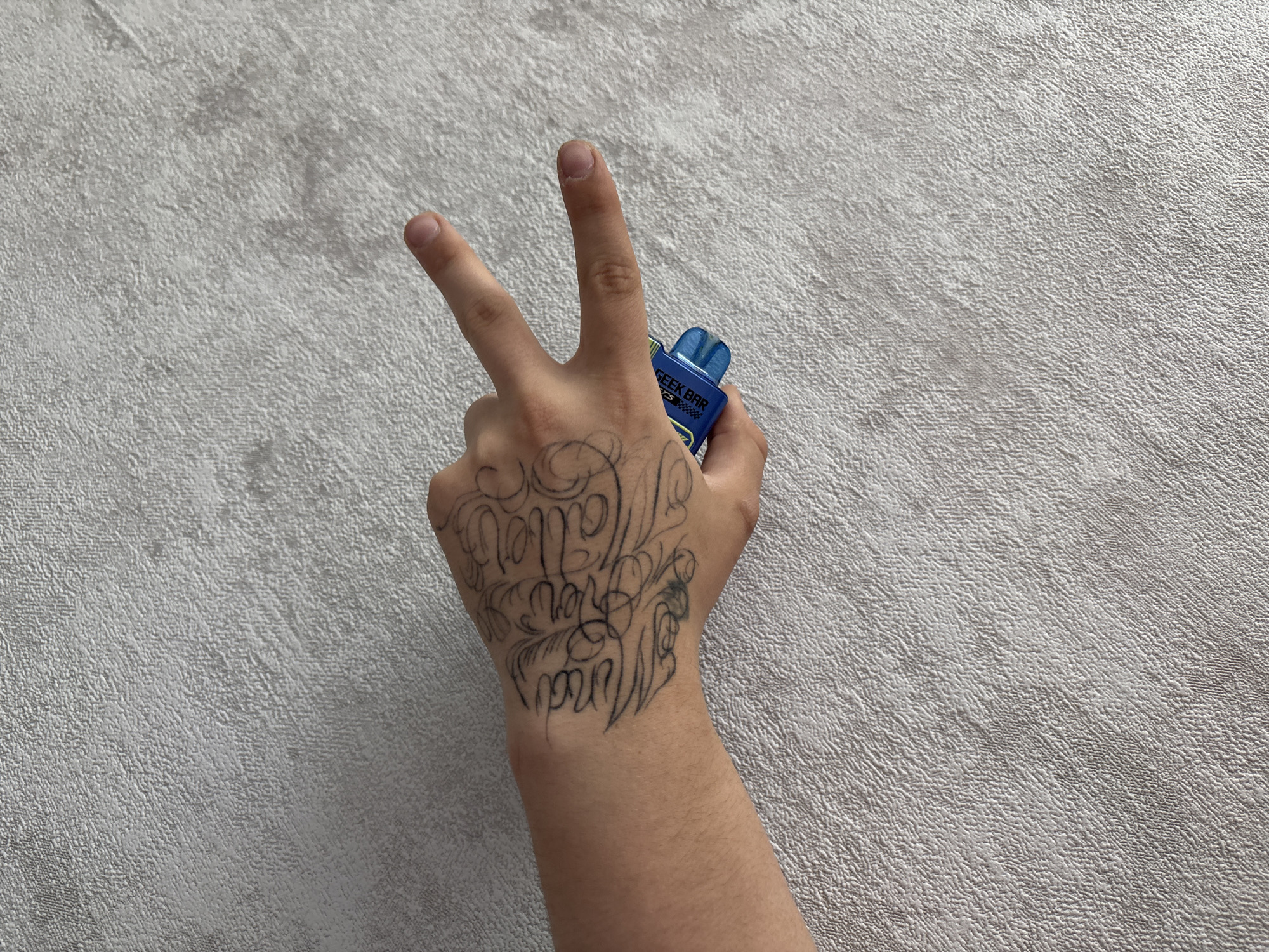 A photograph shows a hand with a tattoo holding a blue Geek Bar vape pen. The hand is in a peace sign gesture, with two fingers extended. The background is a textured gray surface. The location appears to be indoors but is otherwise unknown.