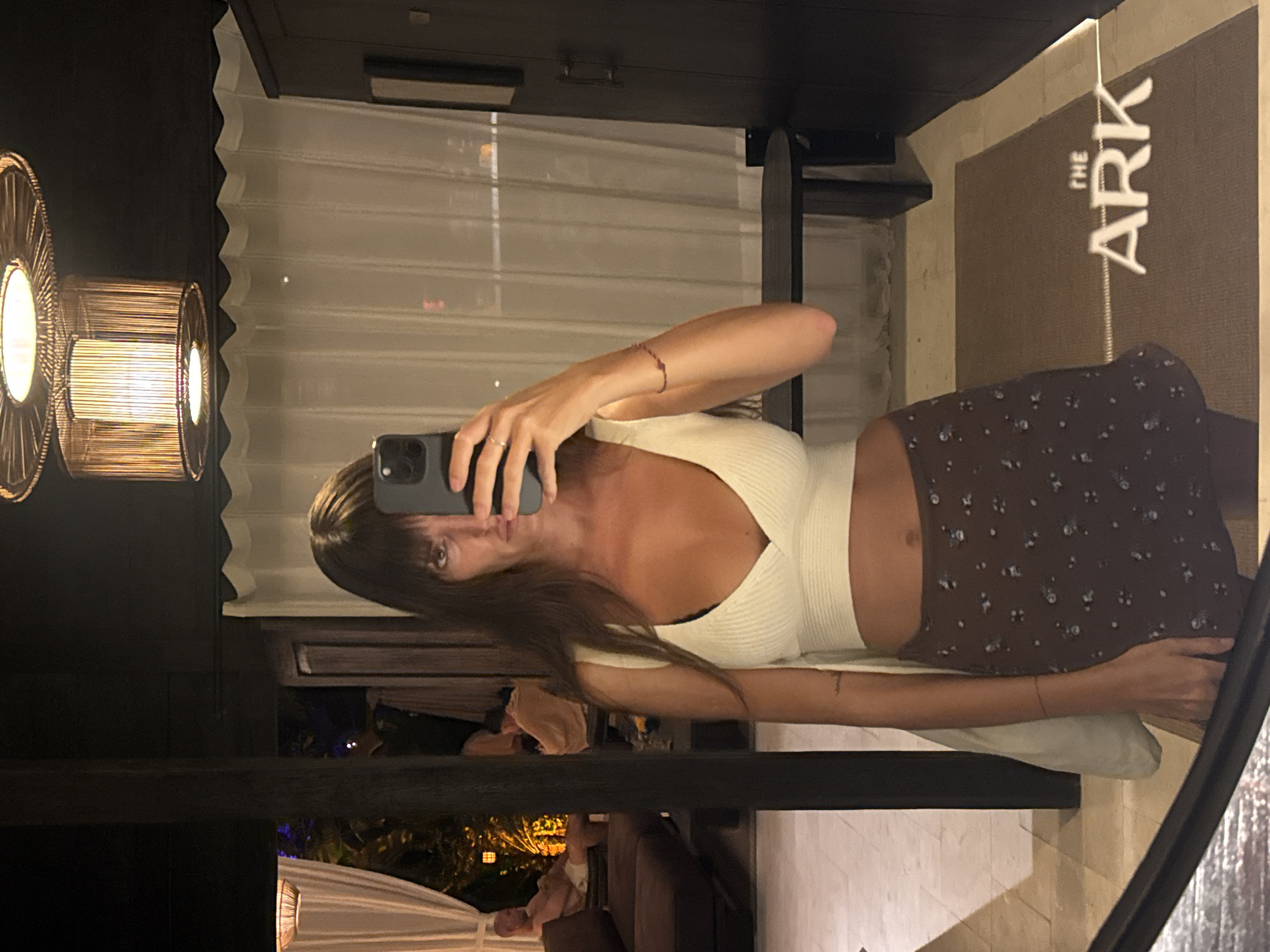 A medium shot shows a fair-skinned, light-haired person standing in a mirror taking a photo of themselves with a phone. The person is wearing a light-colored, ribbed, cropped top and a brown skirt with a floral pattern.
The background reveals an interior setting, likely a room in a hotel, specifically "The Ark" as indicated by the sign visible in the mirror's reflection. The room features decorative lighting fixtures, curtains, and furniture. Outdoor lights and greenery are visible in the background.
The photo was likely taken in Mengwi, Indonesia, given the hotel name reference.