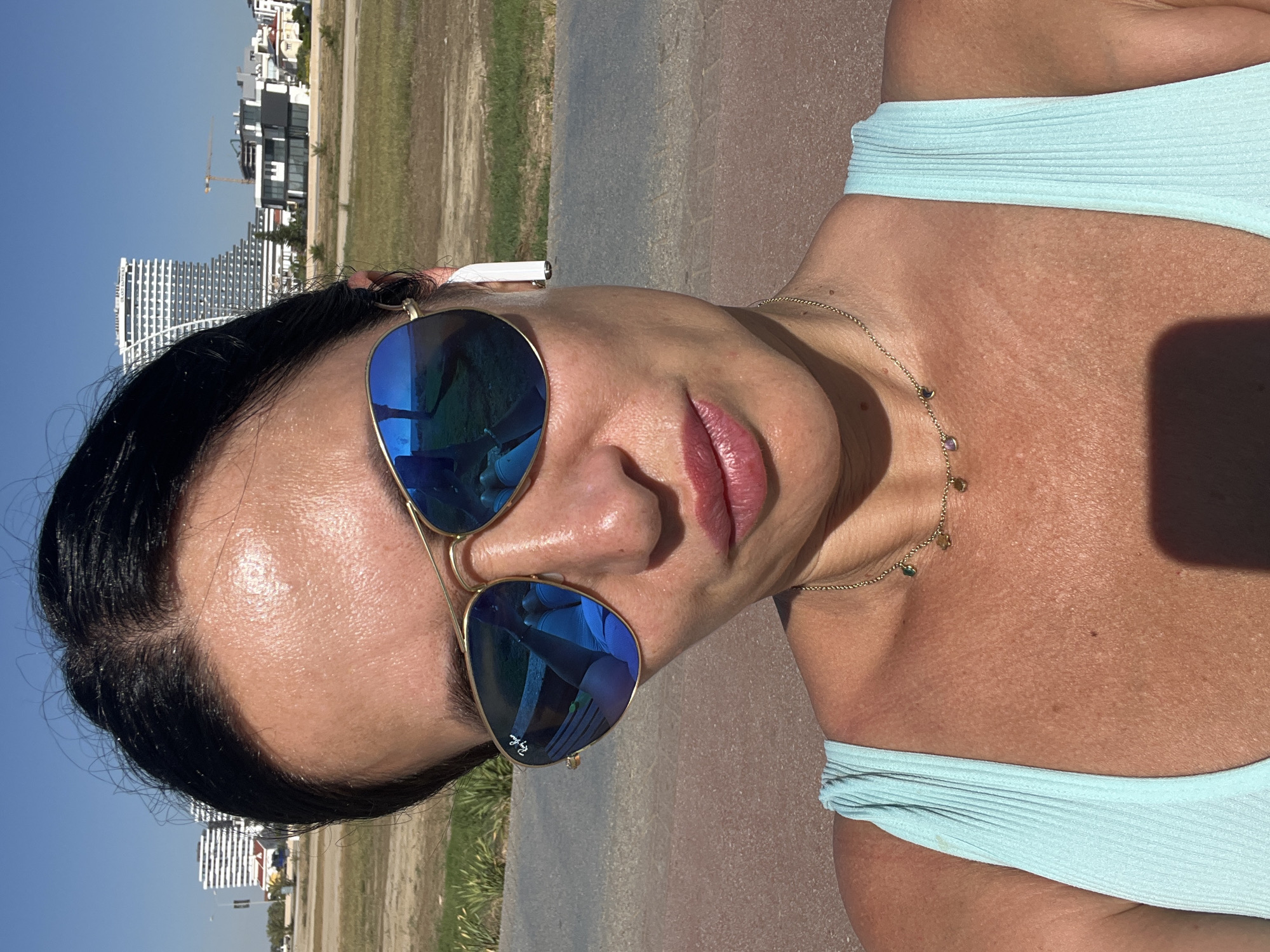 This is a vertically oriented close-up outdoor shot, featuring a woman with dark hair wearing blue mirrored sunglasses and a light blue top or swimsuit. The woman's face takes up a significant portion of the foreground, with her eyes obscured by the reflective lenses of the sunglasses, which bear the "Ray-Ban" logo on the right lens. She has a neutral expression, with her lips slightly parted. A gold chain necklace with small colorful beads is visible around her neck and chest. A white earbud, likely an Apple AirPod, is tucked behind her right ear.
The background shows a sunny, clear blue sky above a landscape with buildings and dry vegetation. On the left side of the frame, several modern buildings, including a tall, white, multi-story structure with a distinctive curved shape, are visible against the sky. A construction crane is also discernible among the buildings. The ground consists of a strip of dry, light brown grass or field, separating the buildings from a darker, paved surface in the immediate foreground and midground, possibly a road or path. The overall impression is a bright, sunny day in an urban or developing area, consistent with the specified location context of Trikomo, Northern Cyprus / Cyprus.