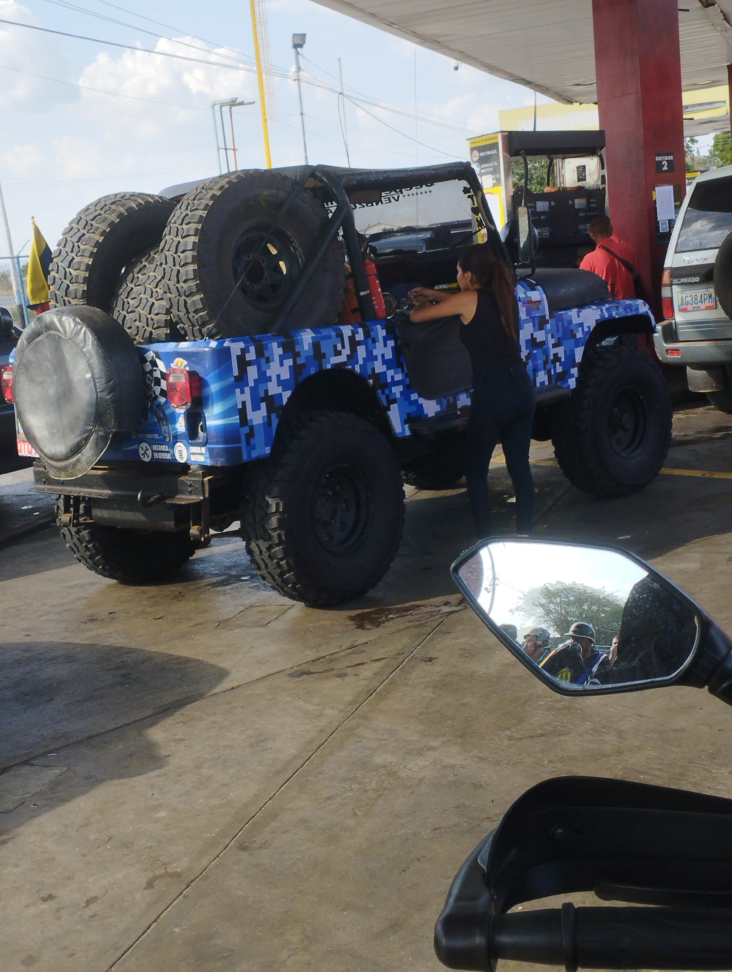 The image shows a gas station in Taguanes, Venezuela. A blue and white pixelated Jeep with large tires is parked next to a pump, with a woman in a black tank top and jeans standing beside it. Another man in a red shirt is near the pump. A silver Toyota Prado with the license plate AG384PM is parked adjacent to the Jeep. A motorcycle's handlebar and a mirror reflecting a group of people are in the foreground.