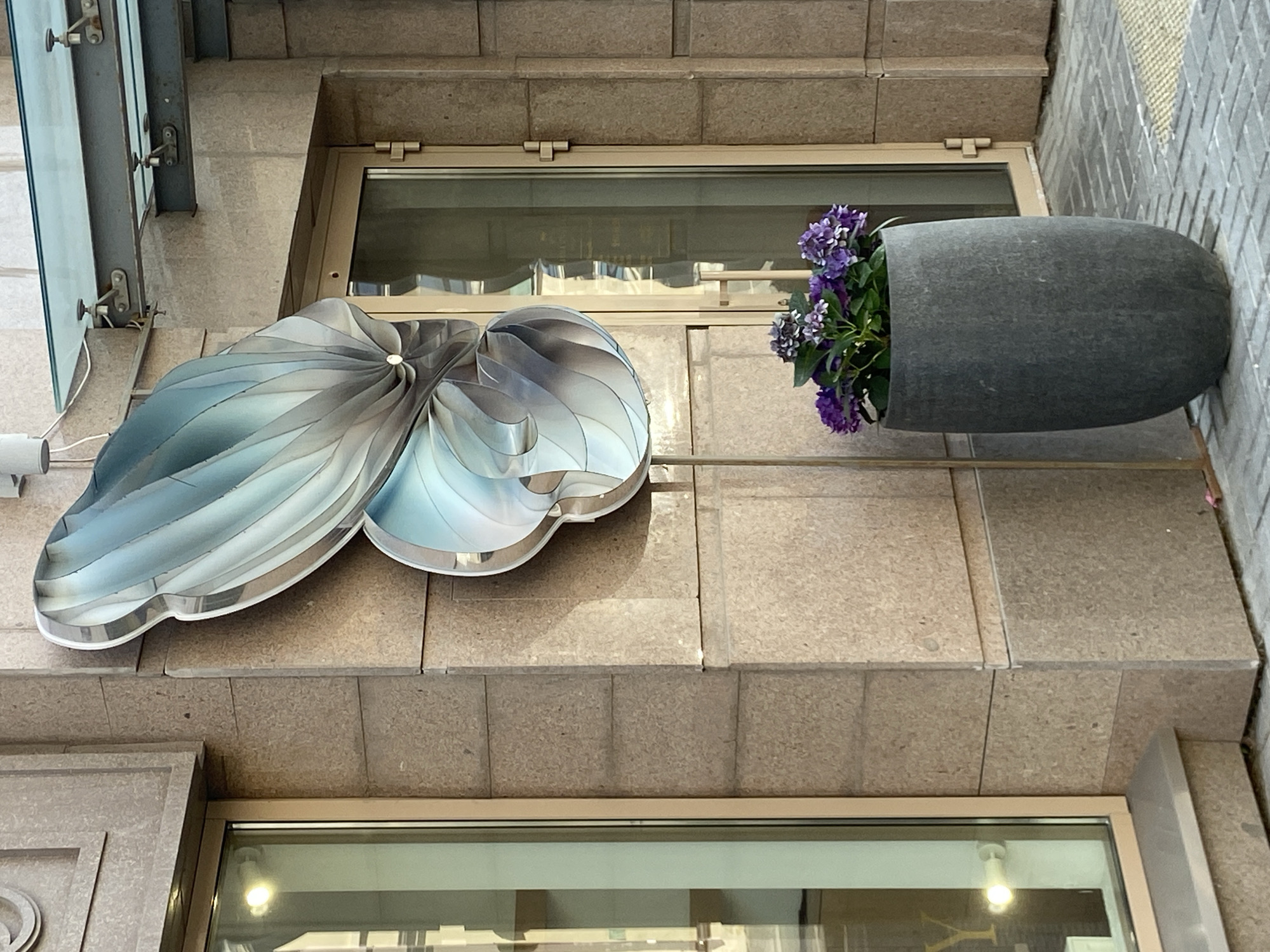 The scene captures an exterior view of a building, likely in Kazan, Russia. The architecture is characterized by light brown stone or tiled surfaces. Two windows are visible, one near the top and another towards the bottom of the frame, both with reflective glass surfaces showing indoor lighting. Attached to the exterior wall is a decorative, reflective blue and gray element, resembling a stylized leaf or swirl. A dark gray, elongated planter with purple flowers is mounted on the wall to the right of the decorative element. The composition is from a low angle, looking upwards towards the building facade.