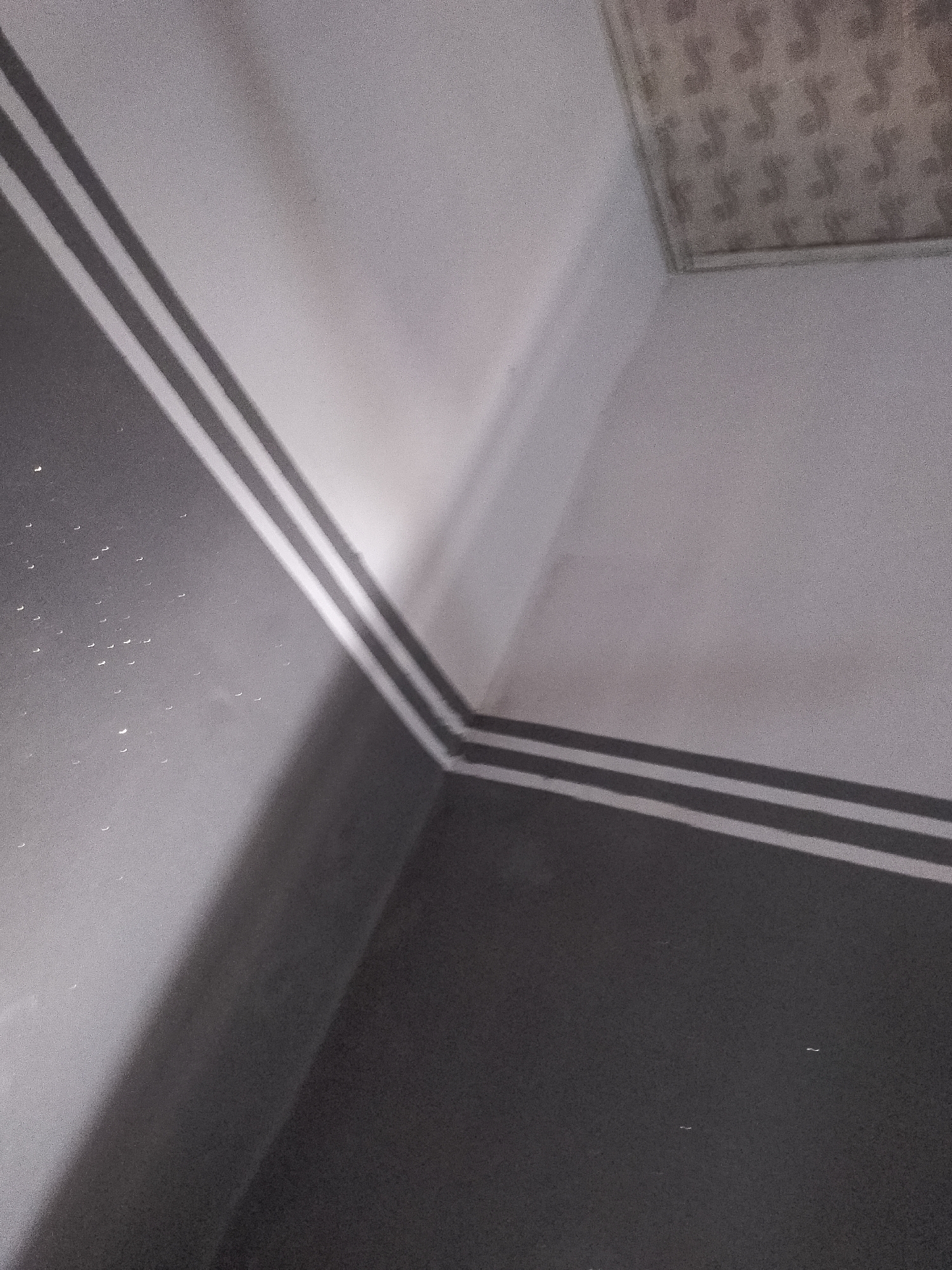 The image shows an interior view of a room, likely a corner where two walls meet. The walls are painted in two distinct colors: a darker shade on the left, transitioning to a lighter shade on the right. A decorative element, appearing as two parallel lines of a contrasting color (possibly white or a lighter gray), runs along the junction of the walls, accentuating the architectural lines. The ceiling is visible in the upper right portion of the image and has a patterned texture. Scattered on the darker wall are small, light-colored specks or reflections, possibly dust or imperfections. The lighting suggests an indoor environment with some light source creating shadows and highlights, emphasizing the textures and colors of the walls. There are no people or specific objects that indicate a particular activity or location within the city of Jalingo, Nigeria, beyond the general description of an interior space.