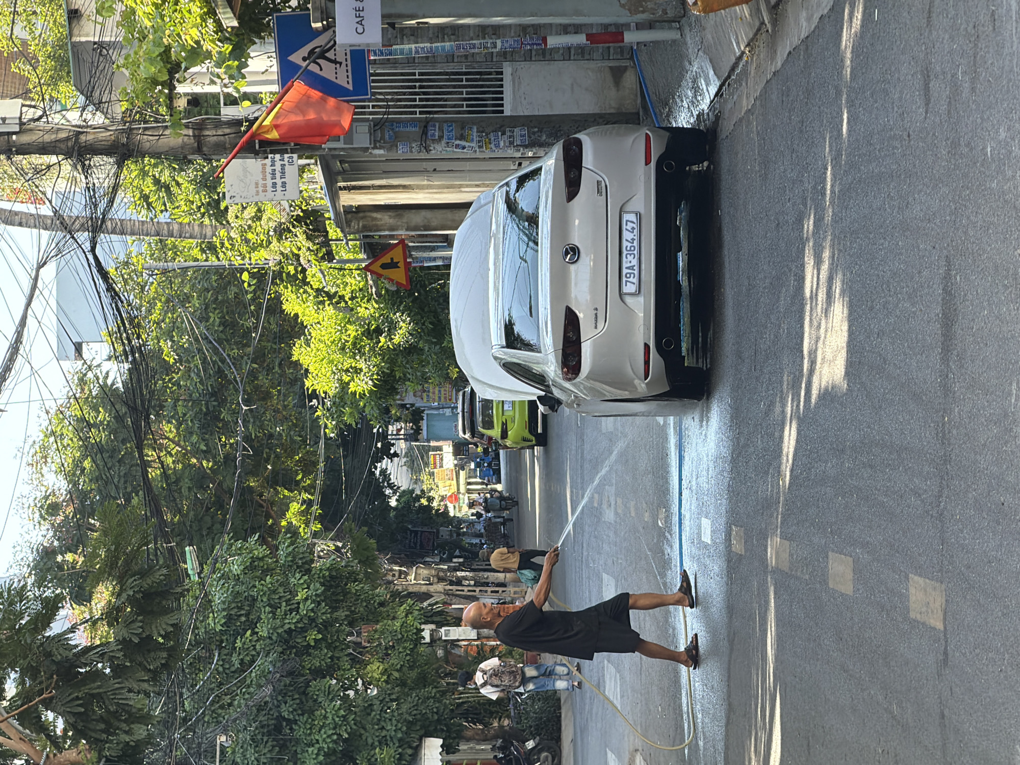 In a street scene in Bac Nha Trang Ward, Vietnam, a bald, middle-aged man wearing a black t-shirt and dark shorts is hosing down the asphalt road. He holds a yellow hose connected to a water source, spraying a stream of water onto the road surface, creating a wet patch. To his right, a light-colored sedan with its hood up is parked on the street. Further down the road, other vehicles and pedestrians are visible, including a lime green car and a person on a scooter. The street is lined with buildings and lush green trees, with a canopy of overhead electrical wires creating a complex network above. A Vietnamese flag is displayed on a pole to the right, alongside street signs and building facades. The overall environment suggests a typical urban street in Vietnam on a sunny day.