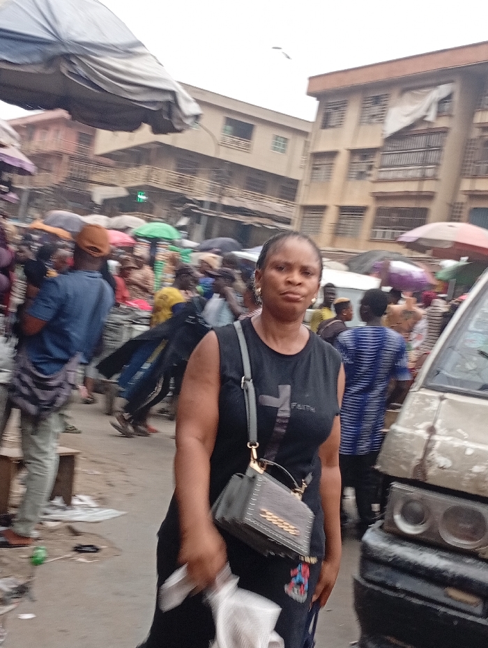 A woman stands in a crowded street scene. She is wearing a black sleeveless shirt with the word "FAITH" and a cross on it, a black skirt, and a gray purse. The street is filled with people and stalls, creating a bustling atmosphere. Buildings rise in the background, adding to the urban setting. A vehicle is seen partially on the right, suggesting the presence of traffic. The scene appears to be taking place in a busy market or commercial area.