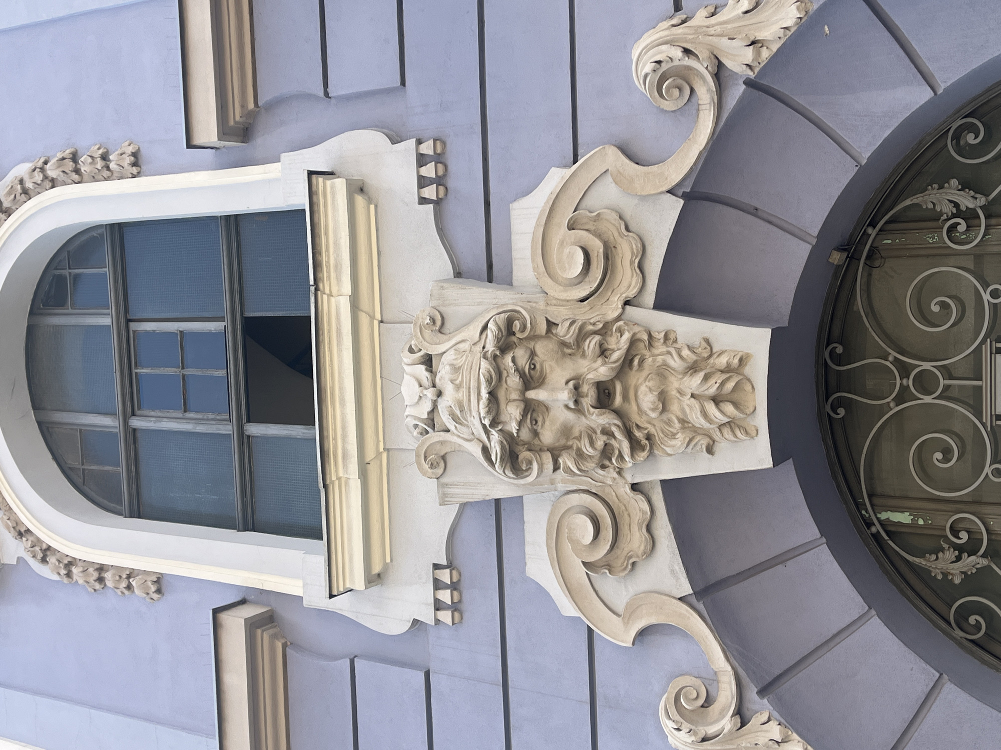 This is a close-up, eye-level, outdoor, daylight shot of architectural ornamentation above a doorway. The facade of the building is painted a light purple color. The central feature is a stone carving of a male face with a beard and a helmet. The carving is elaborately decorated with swirling decorative elements on either side. Above the carving is a large arched window with multiple panes. The window is partially open. The overall style suggests Baroque or Neoclassical architecture. The scene is static and there are no people visible.