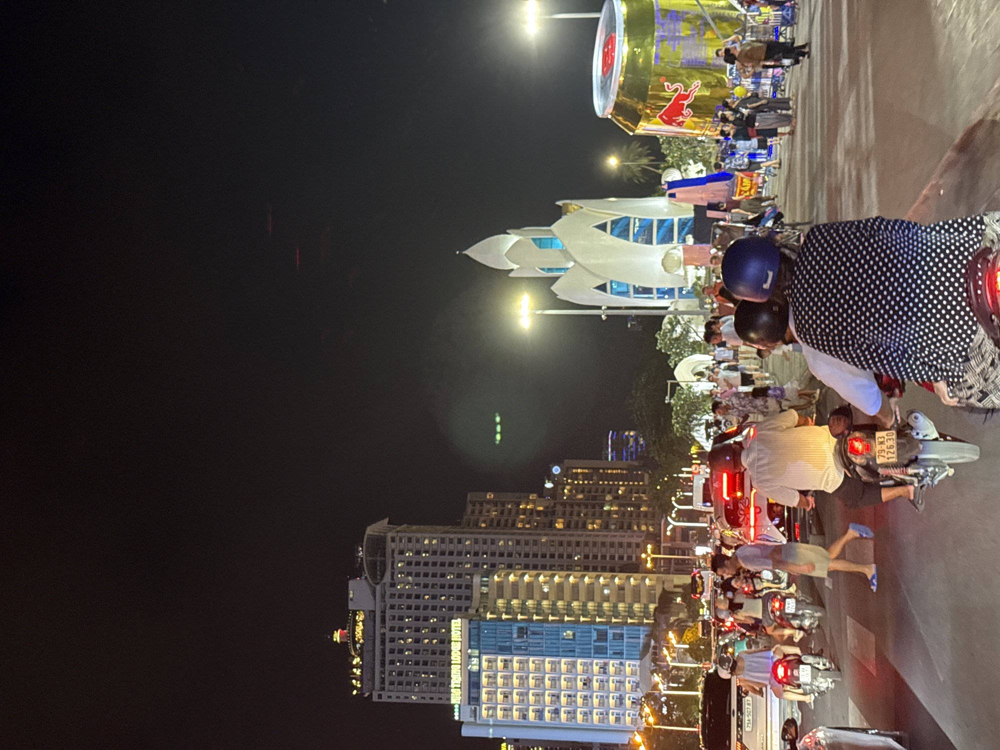 This nighttime street scene in Nha Trang, Vietnam, depicts a busy urban environment, likely a thoroughfare with heavy traffic. The primary subjects are numerous people on and around motorcycles, suggesting a common mode of transportation in the area. The atmosphere is lively, with people dressed in casual clothing, some wearing helmets. The time of day is clearly night, evidenced by the dark sky and the illuminated buildings and streetlights.
Notable objects include several motorcycles, some parked and others in motion, contributing to the congested feel of the scene. A large, golden Red Bull can advertisement is prominently displayed in the upper portion of the image, indicating a commercial or promotional area. The architecture of the buildings suggests a modern city, with tall structures featuring many illuminated windows. One building in the foreground has the text "THE HOIAN LODGE HOTEL" visible.
The weather appears clear, allowing for good visibility despite the darkness. The overall mood is one of bustling activity and the vibrant energy of a city at night. The scene captures the dynamic street life of Nha Trang, with a focus on the constant flow of traffic and pedestrian activity.