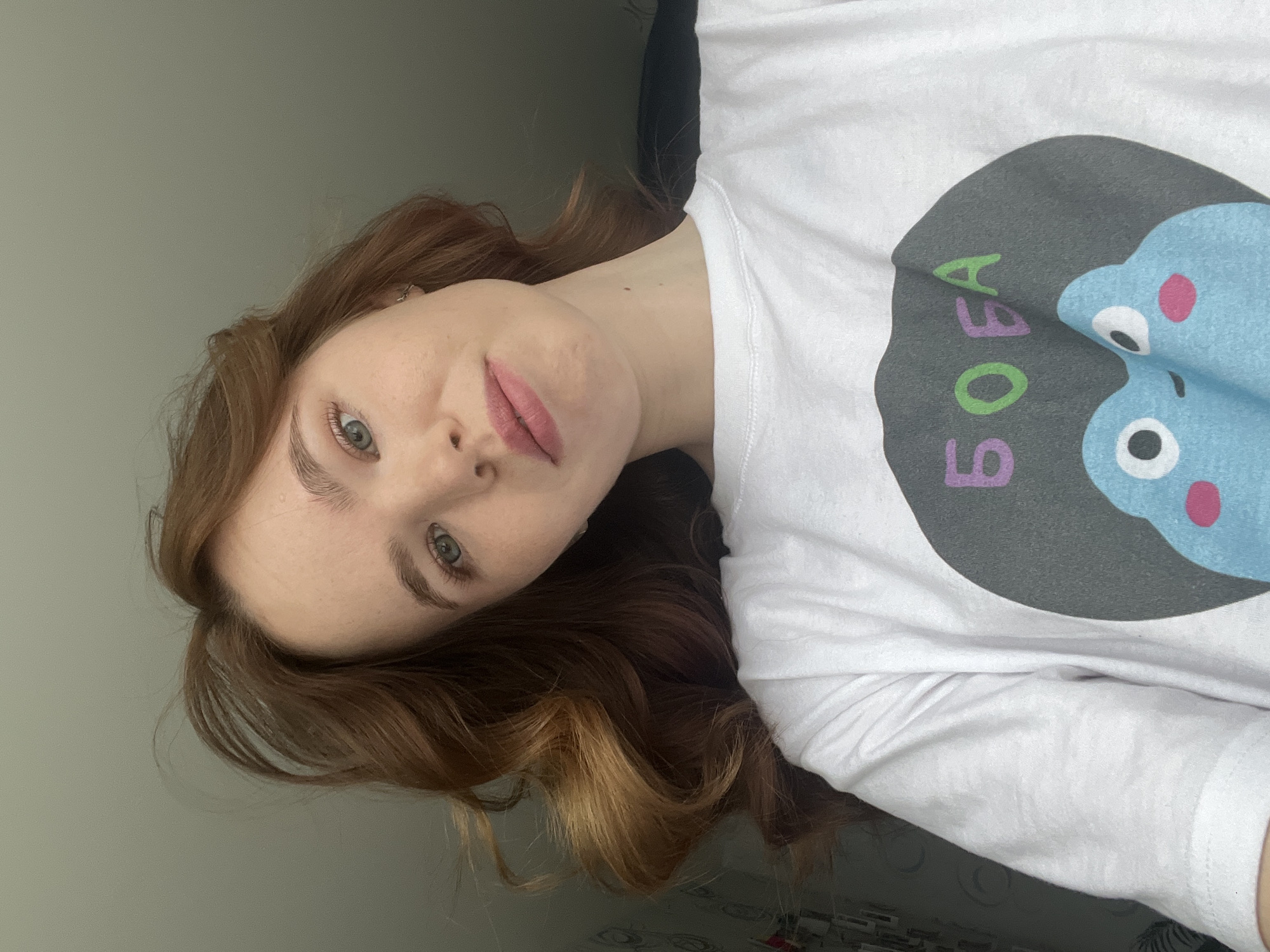 A young woman with auburn hair and fair skin poses indoors. She wears a white t-shirt printed with a cartoon image and the Russian word "БОБА" in purple and green letters. Her head is tilted, and she is looking directly at the camera. The background is a plain, light grey wall. The location is Chaikovsky, Russia.