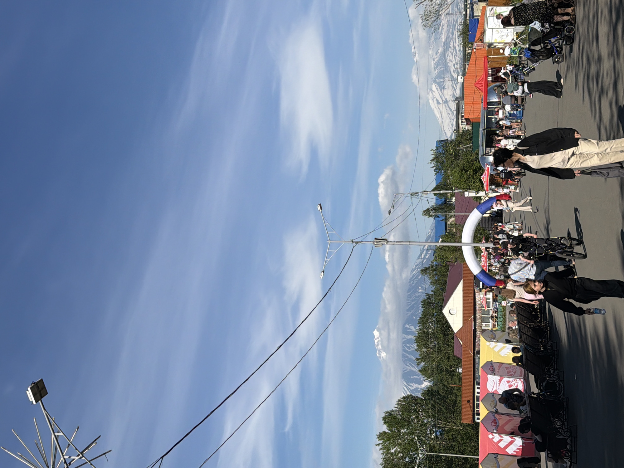In Yelizovo, Russia, on a bright, clear day, a street fair is in full swing. The foreground is dominated by a paved road where people are walking. In the mid-ground, a row of colorful tents, likely stalls or booths, are set up along the side of the road, with people browsing or sitting in chairs in front of them. An inflatable archway, adorned with red and blue elements, stands prominently, marking an entrance or a point of interest. Beyond the tents and the crowd, buildings of varying colors and designs are visible, some with red roofs. In the background, majestic, snow-capped mountains rise against a vivid blue sky dotted with wispy clouds. The scene is bustling with activity, with individuals walking, pushing strollers, and riding bicycles, suggesting a lively community event.