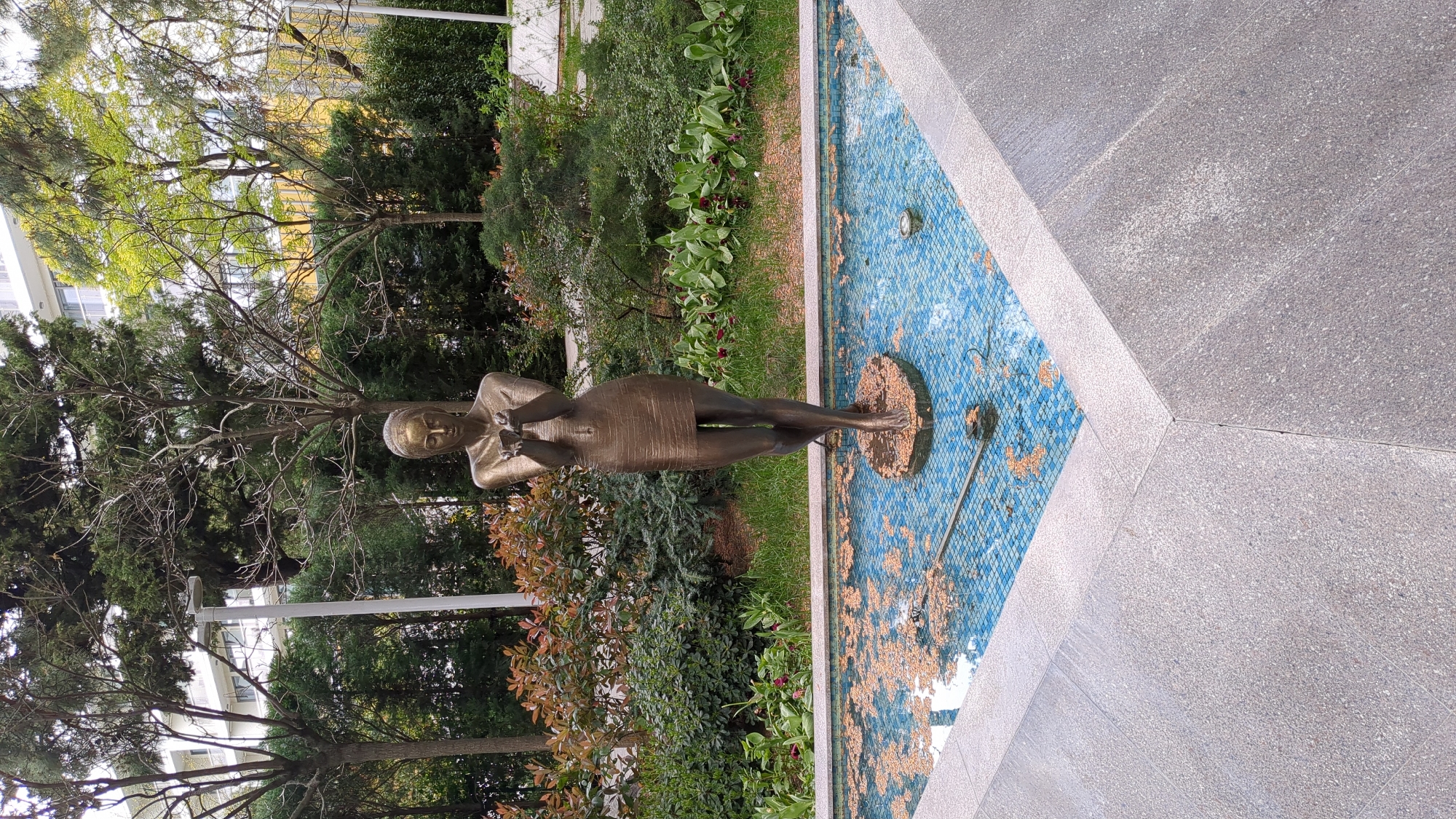 A bronze statue of a woman stands in a blue-tiled fountain. The fountain is edged with grey concrete and surrounded by trees and greenery. The location is Gelendzhik, Russia.