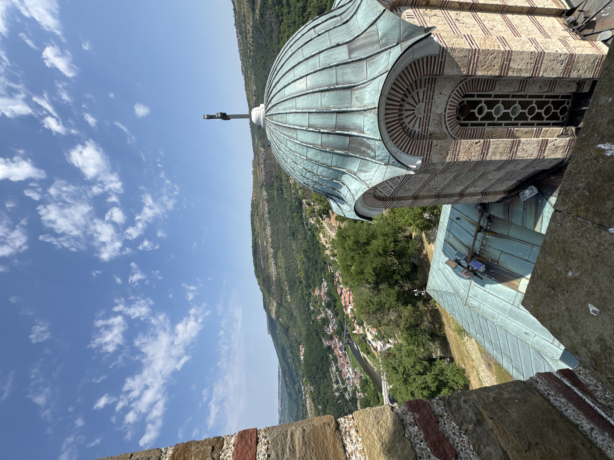 The image captures a view from a stone structure, likely a castle or fortification, overlooking the city of Veliko Tarnovo, Bulgaria. In the foreground, a portion of a stone wall with alternating red brick and light stone courses is visible on the left. To the right, the curved, oxidized copper dome of a church or chapel is prominent, topped with a cross. The dome is supported by a multi-story structure with decorative brickwork and a tall, narrow window featuring a circular pattern. Adjacent to the dome is a sloped, weathered copper roof.
Beyond these architectural elements, the scene opens up to a panoramic vista of Veliko Tarnovo. A winding river, possibly the Yantra River, flows through the valley, crossed by bridges. The city itself is nestled within the valley, characterized by terracotta-tiled roofs and scattered buildings. The surrounding landscape is a verdant tapestry of rolling hills covered in dense green forests. The sky above is a clear blue, dotted with scattered white cumulus clouds. The overall impression is one of historical significance and natural beauty.