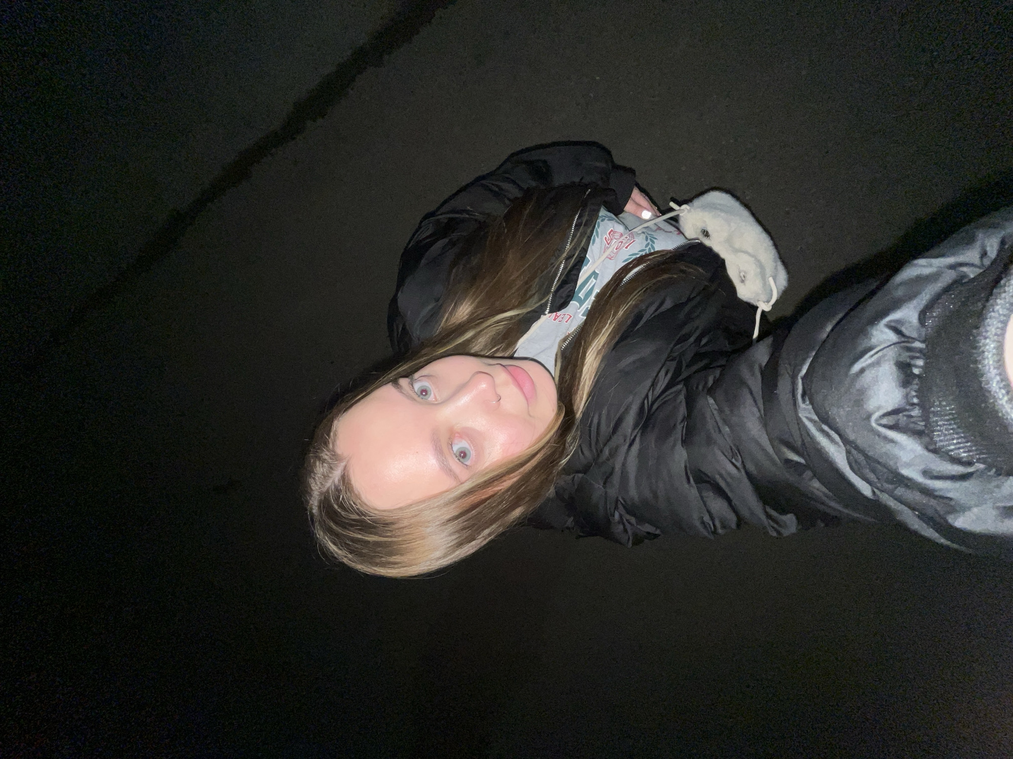 A young woman with blonde hair and light blue eyes is standing outside at night. She is wearing a black puffer jacket over a light grey sweatshirt. She is holding a light-colored, fluffy purse with drawstring closures. The environment is dark, possibly a paved road or street. The location is unknown, Novopokrovskaya is not discernable in the image.