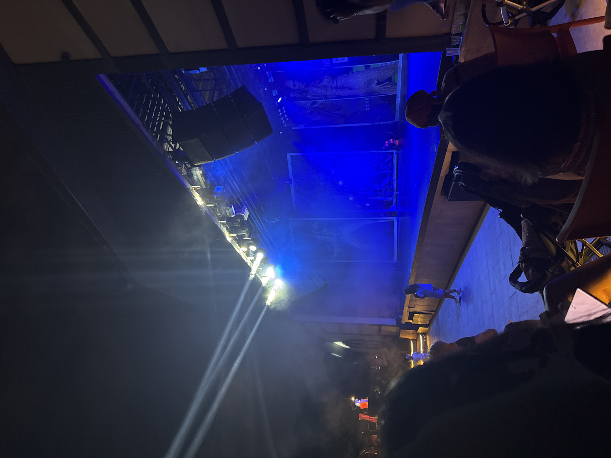 The image shows an outdoor evening event at a venue in Aksu, Turkey. The scene is illuminated by stage lighting, with bright white beams cutting through a smoky atmosphere from the left. A stage is visible in the background, lit with a vibrant blue light. Large speakers are suspended above the stage. On the stage, there are large posters or backdrops depicting figures. Several people are present in the foreground, mostly silhouetted against the stage lights, suggesting they are an audience. Some are seated, while others appear to be standing or walking. Two young children are also visible near the stage. The overall setting appears to be an entertainment venue, possibly a theater or an open-air concert space.
