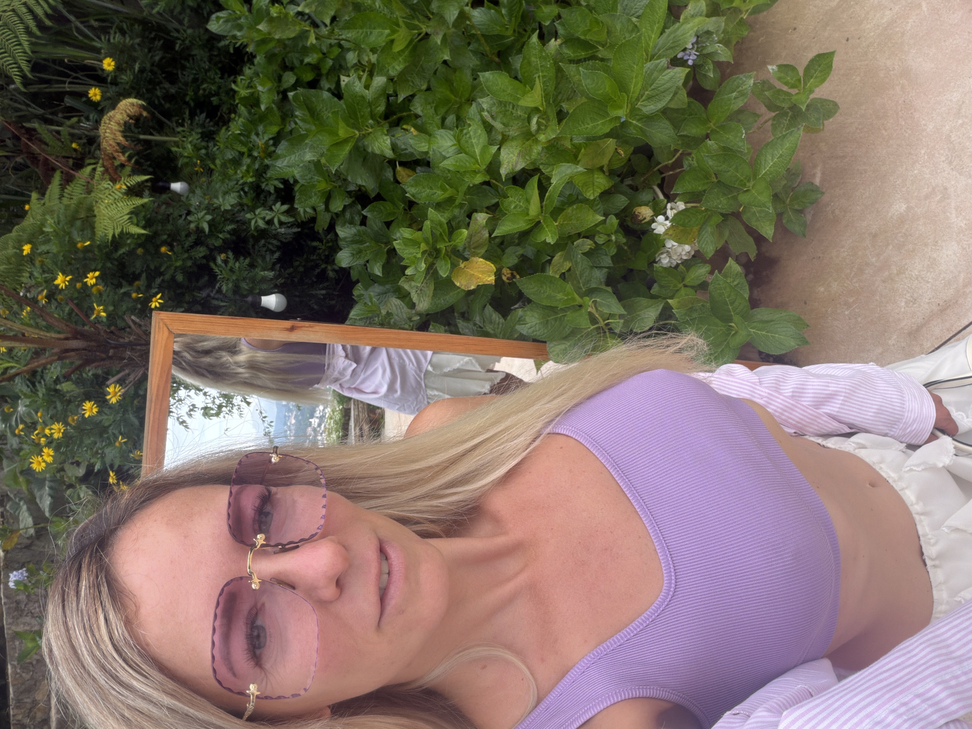 A woman with blonde hair and pink-tinted sunglasses is taking a selfie in front of a mirror, which is propped up outdoors. She is wearing a light purple, ribbed crop top and a white striped shirt over it. Behind her, there is lush green foliage, some yellow flowers, and a hint of a cityscape in the background, suggesting she is in Xuân Thành, Vietnam. The mirror reflects her back, showing her in a light purple dress. The overall scene is bright and summery.