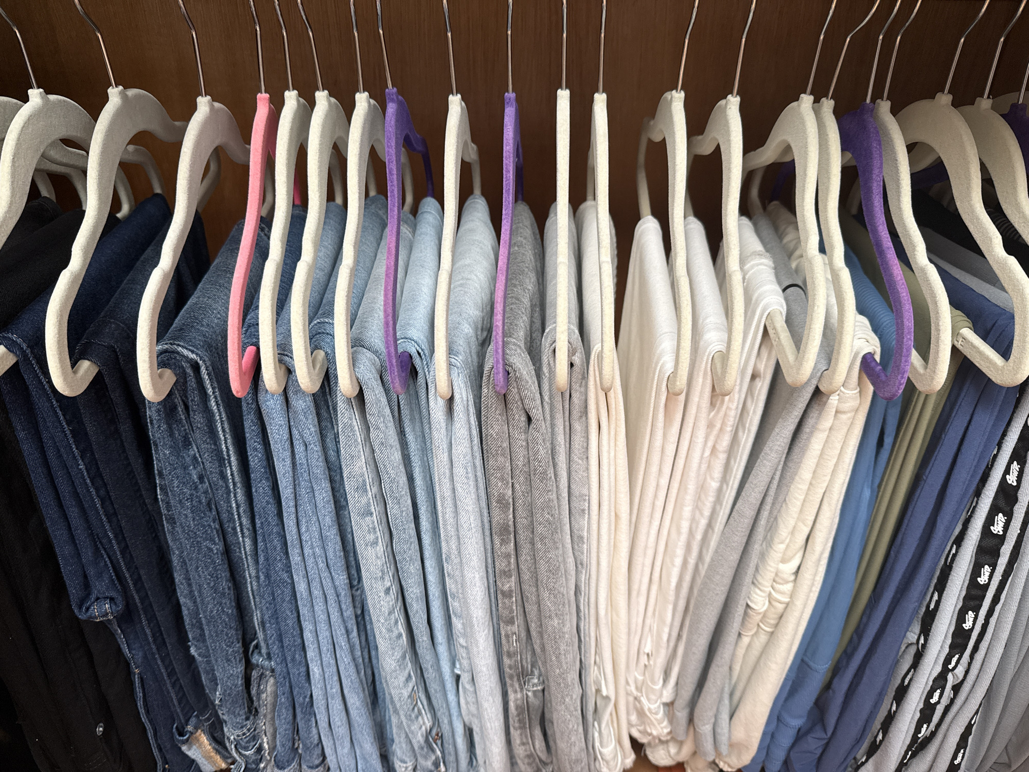 The image shows a close-up, eye-level view of a closet filled with a variety of pants hanging on hangers. The hangers are mostly off-white, with a few pink and purple hangers interspersed. The pants range in color from dark denim to light gray and white. The background appears to be the wooden interior of the closet. No people are visible. The location of San Bernardino Tlaxcalancingo, Mexico, is not discernible from the image.