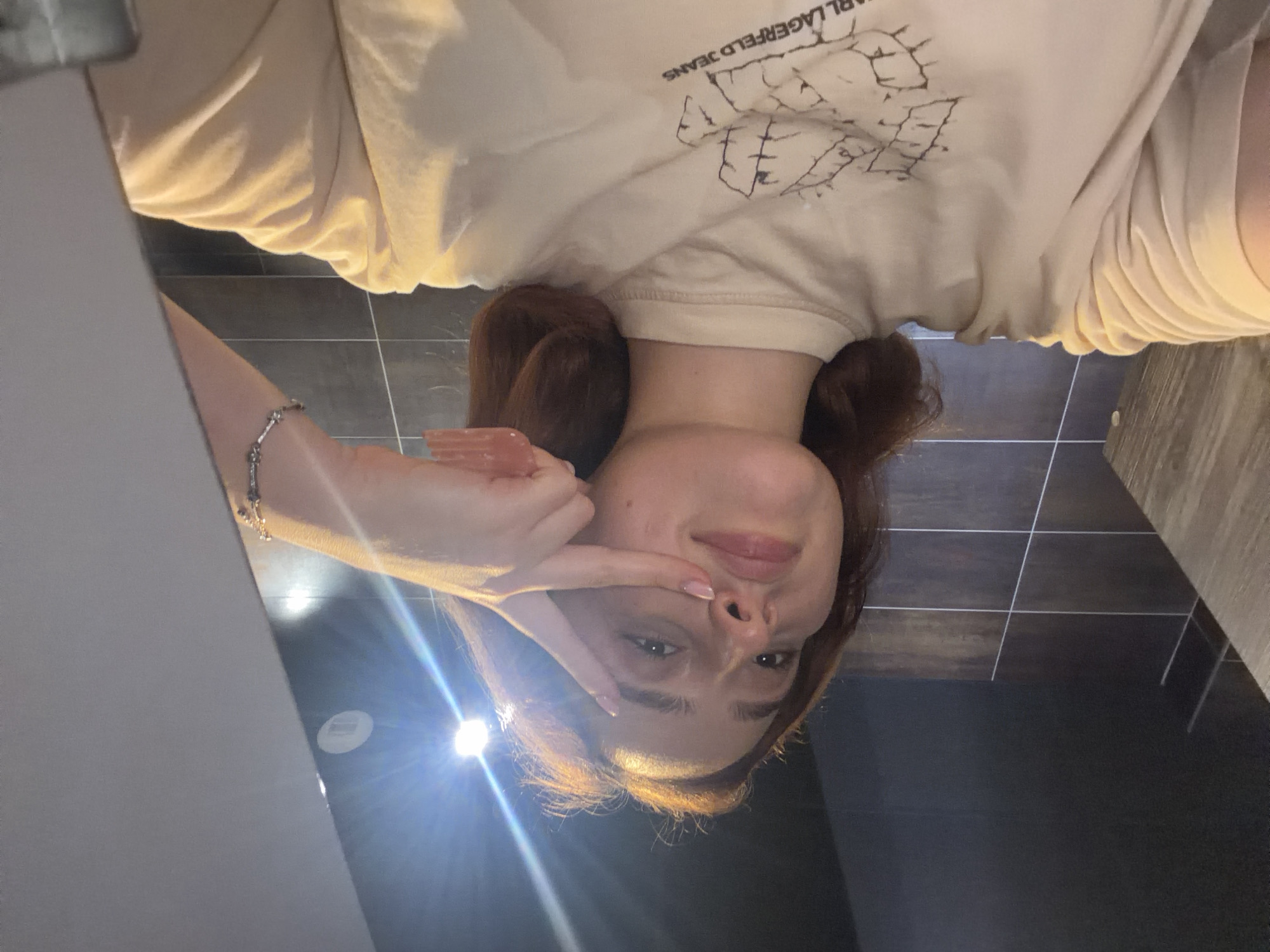 A young woman with reddish-brown hair is taking a selfie in what appears to be a public restroom. She is wearing a light beige t-shirt with a graphic print that includes the text "KARL LAGERFELD JEANS". Her left hand is raised, with her index finger and middle finger extended in a peace sign gesture, touching her cheek. A bracelet with small charms is visible on her right wrist. The background shows tiled walls and dark ceiling panels, with bright overhead lighting creating reflections. The image does not provide any information about the city of Strasbourg, France.