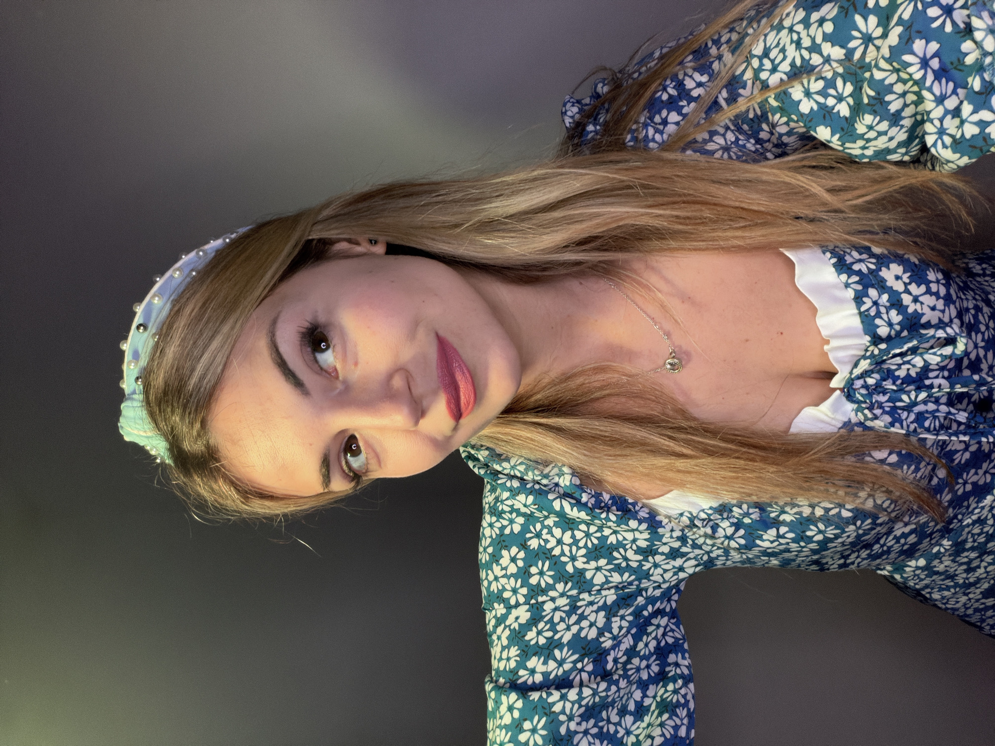 A young woman with long blonde hair is shown in a close-up selfie-style portrait. She is wearing a blue and white floral print dress with a white ruffled neckline, and a light blue headband adorned with small pearls. A silver necklace is visible around her neck. The background is a gradient of gray, fading from lighter to darker. The location in the city of Budva, Montenegro is indeterminable.
