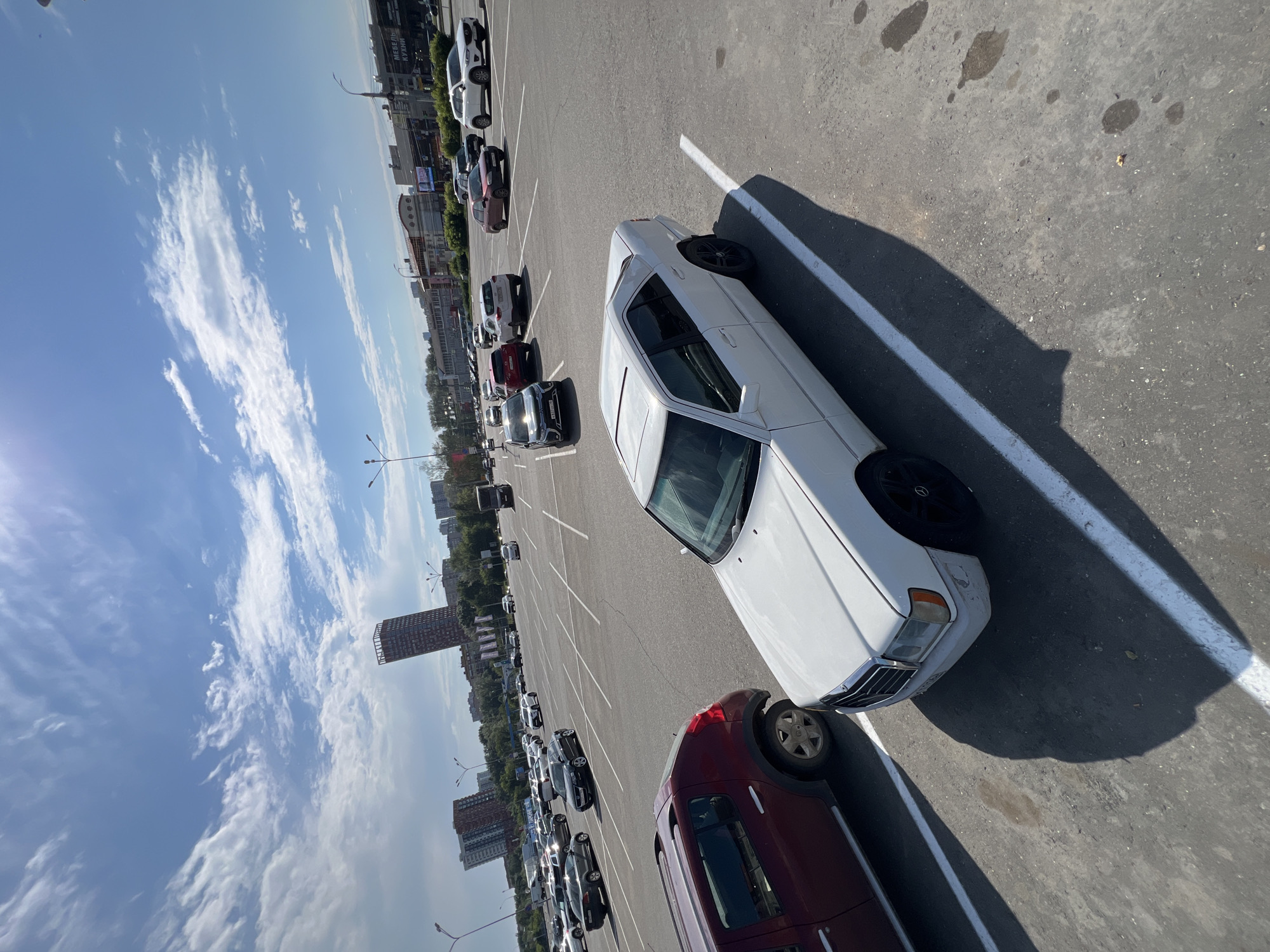 The image shows a large, mostly empty outdoor parking lot under a partly cloudy blue sky. In the foreground, a white sedan is parked diagonally across two parking spaces. To the left of the sedan, a dark red SUV is parked in a single space. The parking lot is paved with asphalt and marked with white lines. In the background, several other cars are visible, along with buildings and trees. The context provided indicates this scene is located in the city of Izhevsk, Russia. The buildings in the background include a tall, distinct reddish-brown tower with a lattice-like structure at its top, characteristic of some Soviet-era architecture, and other mid-rise apartment buildings and commercial structures. Streetlights are also visible in the distance. There are no people visible in the image. The overall impression is of a typical urban parking area on a clear day.