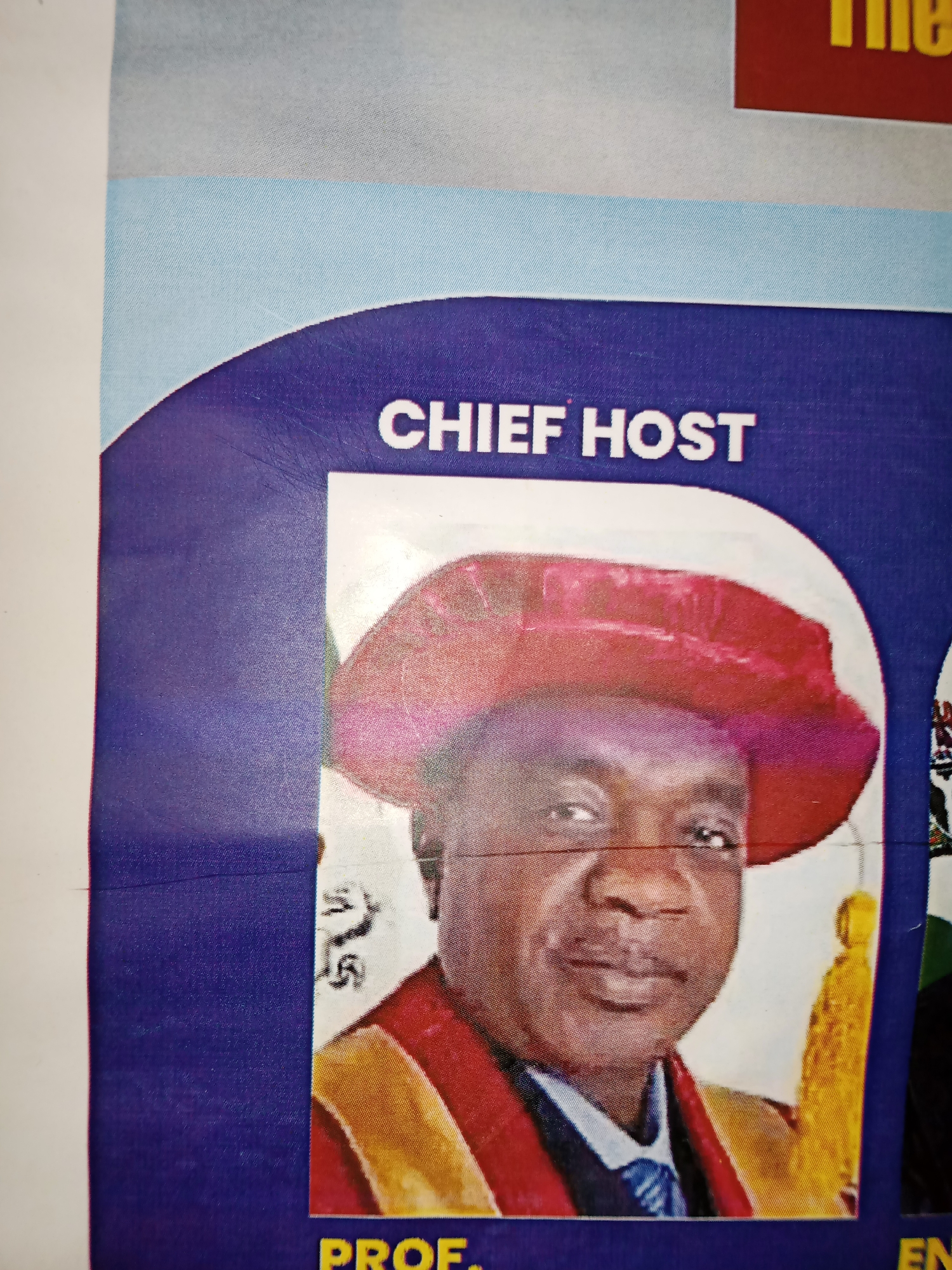 This is a close-up, indoor, eye-level, static photograph of a poster or banner featuring a man in academic regalia. The man, who appears to be of African descent, is wearing a dark cap with a red band and a red academic gown with gold trim over a blue and white collared shirt and tie. He has a mustache and is looking towards the camera with a slight smile. The poster has text that reads "CHIEF HOST" in large white capital letters at the top. Below the man's image, the text "PROF." is visible in yellow capital letters. The background of the poster is a gradient of light blue to white at the top, and a dark purple curved design framing the man's portrait. The image is taken at an angle, suggesting it is part of a larger display. The lighting appears to be artificial and somewhat diffused. Without further context or visible landmarks, the specific location in Jalingo, Nigeria, cannot be determined from the image alone.