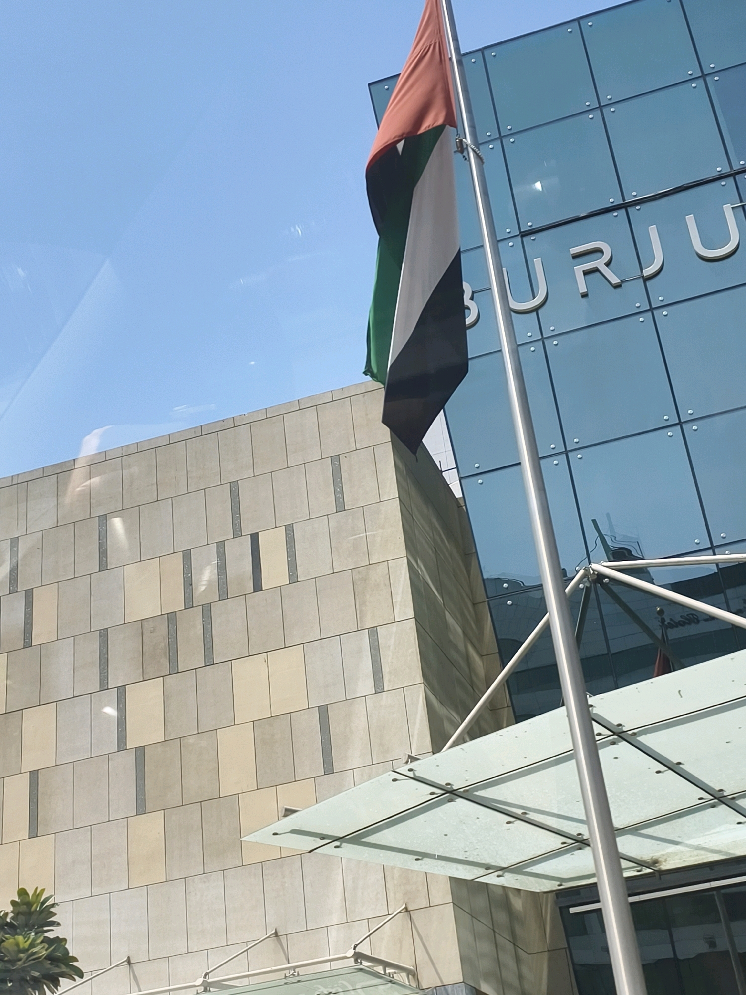 The photograph captures a low-angle view of a modern building exterior in Dubai, United Arab Emirates. A flag, predominantly featuring green, white, and black vertical stripes with a red vertical stripe on the hoist side, flies from a metal flagpole. The building behind the flag is characterized by a glass facade with a grid pattern of metal frames, reflecting the sky and surrounding structures. Silver lettering spelling out "BURJU" is visible on the glass facade. To the left of the glass building, a section of a beige stone-clad wall is present, laid in a brick pattern with darker vertical accents. A clear glass canopy with metal supports extends from the building. The sky is blue and clear, indicating daylight. A small, leafy green plant is visible in the lower-left corner of the frame. The scene suggests a corporate or commercial setting, possibly near a prominent landmark given the "BURJU" lettering, which could indicate proximity to the Burj Al Arab or similar structures.