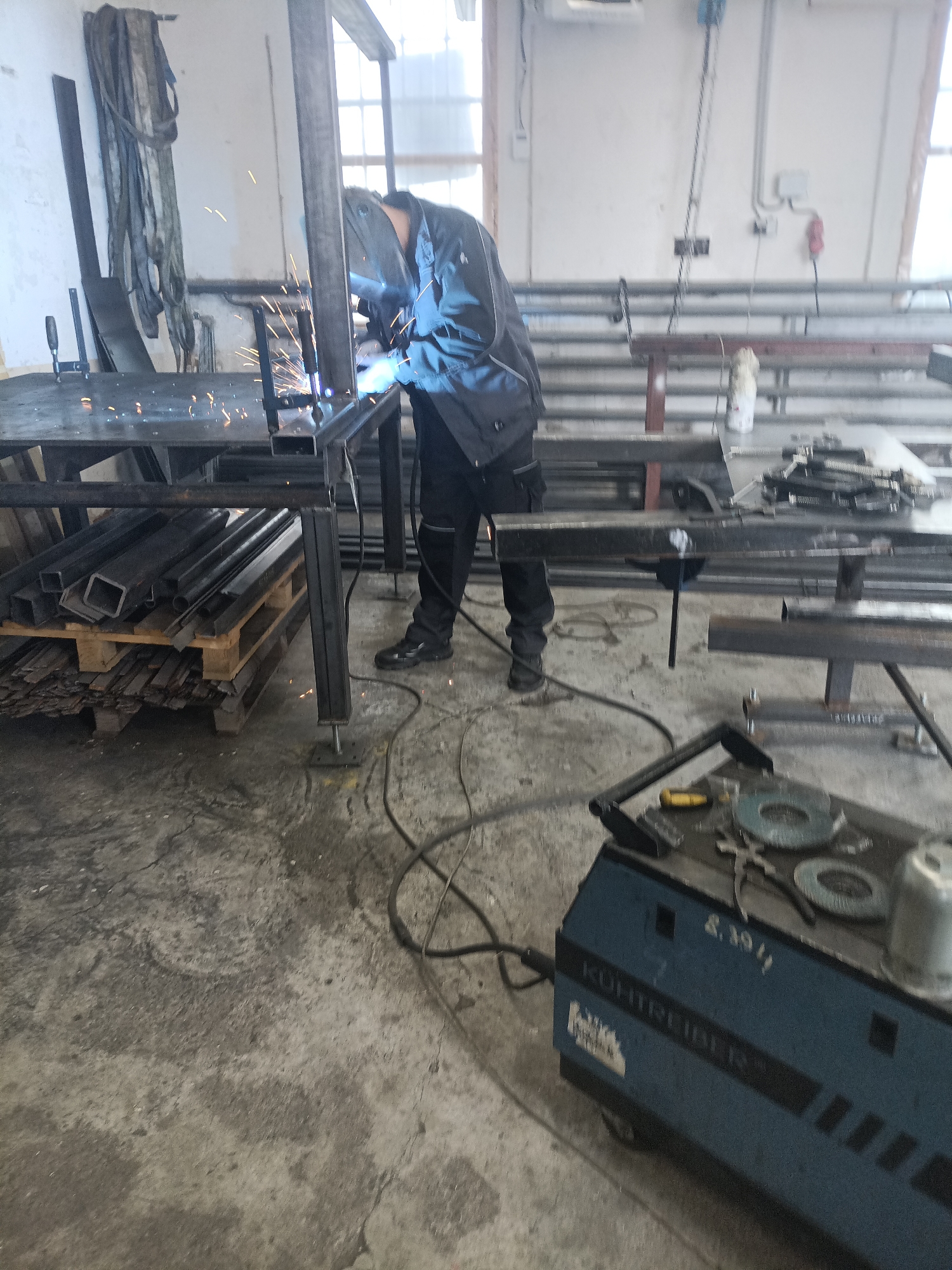 The image depicts an industrial workshop in Dalovy, Czechia, during what appears to be daytime, with natural light entering from windows. The primary subject is a person, whose face is obscured by a welding mask, actively welding a metal structure. Sparks are clearly visible emanating from the point of contact, along with a bright light from the welding arc. The individual is dressed in dark work attire and protective footwear.
They are working on a metal frame, possibly a table or a component of a larger structure, which rests on a robust work table made of dark metal profiles. This work table is supported by adjustable feet. Beneath the table, a stock of metal materials, including square tubing and flat bars, is neatly stacked on wooden pallets.
In the foreground on the right, a blue and black welding machine is positioned on the concrete floor. The brand name "KÜHTREIBER" is visible on its side, along with the partially visible numbers "2.39/1". Various tools, such as grinding discs, pliers, and a yellow-handled object, are placed on top of the machine. Power cables from the welder extend across the concrete floor towards the work area.
The background features a light-colored wall with industrial elements, including horizontal pipes, electrical outlets (one red), and long, dark straps hanging down. The concrete floor shows signs of heavy use, consistent with a workshop environment. Other metal components and structures are scattered throughout the scene, suggesting a busy fabrication setting.