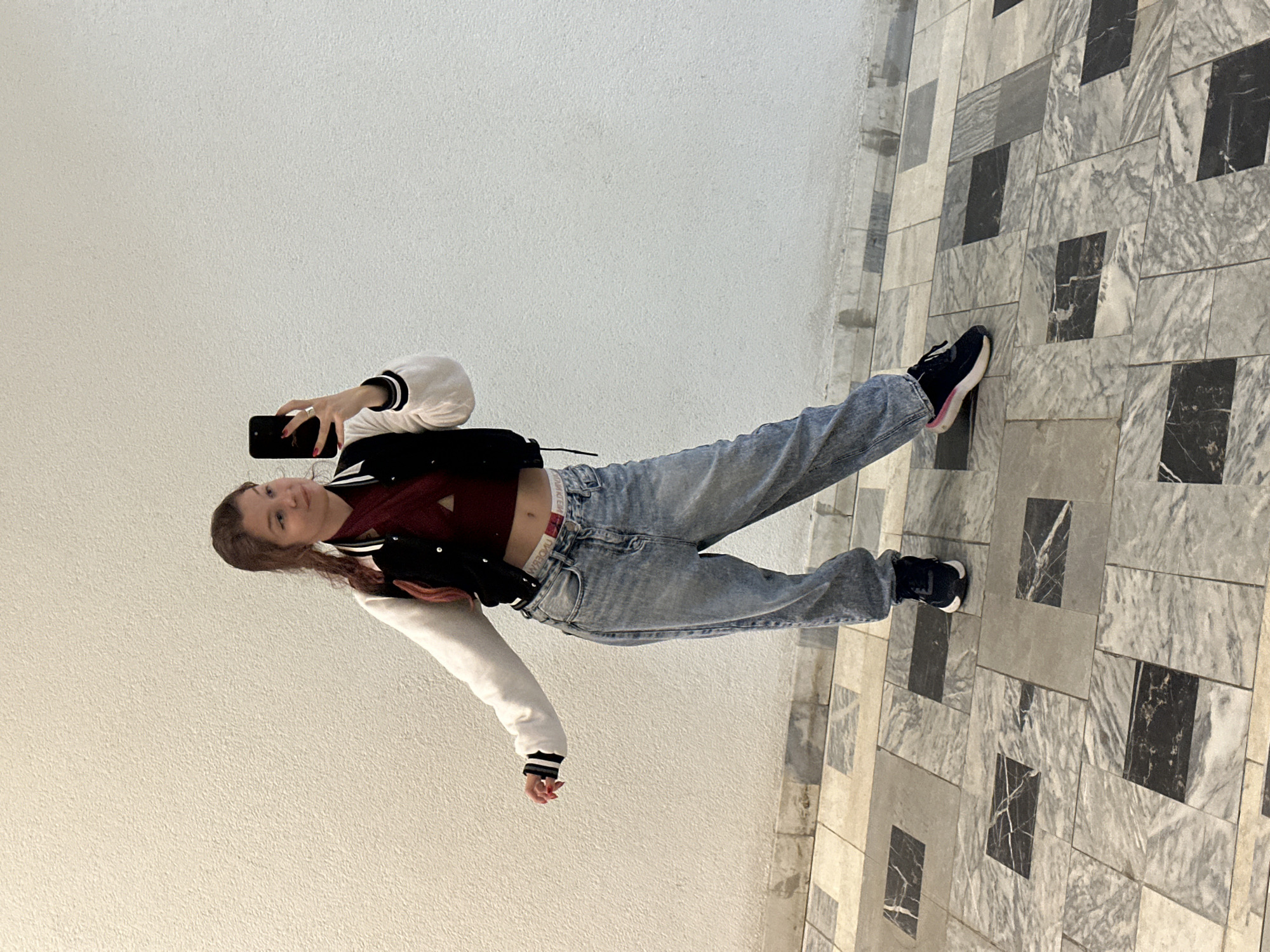 In an indoor hallway with a textured off-white wall and a marble tile floor, a young woman with auburn hair in a ponytail is taking a selfie with her smartphone. She is wearing a maroon crop top, a black and white varsity jacket, and loose-fitting light-wash jeans. She has a ring on her right ring finger. Her left arm is outstretched, and her right arm is holding the phone up. The setting appears to be a modern building, possibly a shopping mall or public institution, given the polished floors and smooth wall.