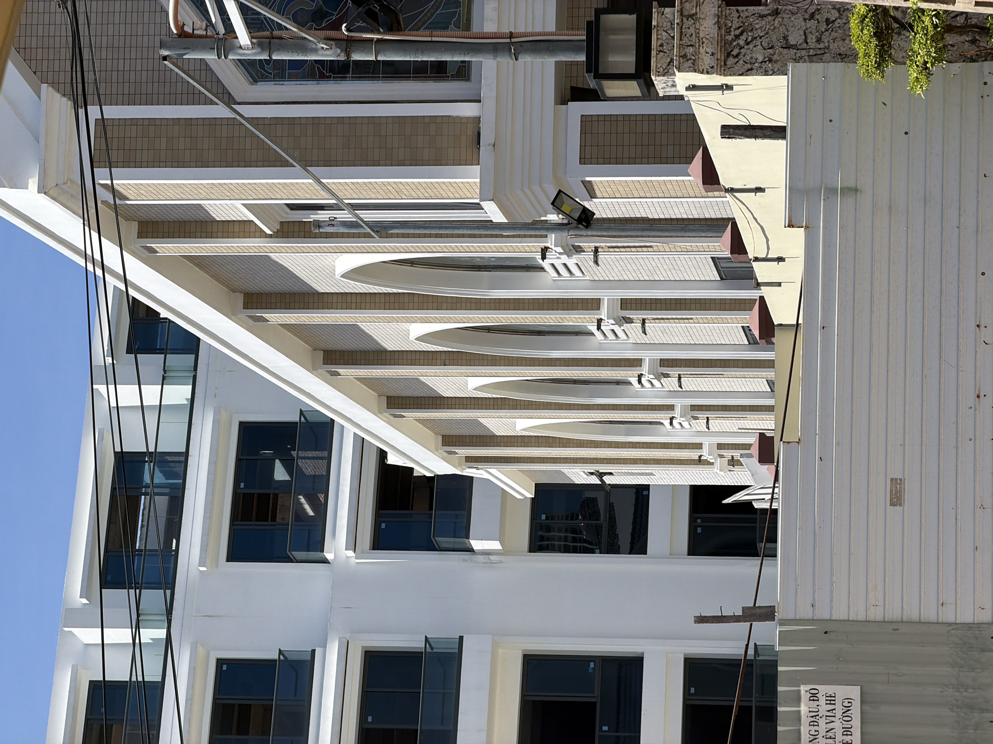 A low-angle view of a building in Nha Trang Ward, Vietnam, presents a modern architectural design featuring repetitive arched awnings that run along the façade. The building is primarily white with dark-framed windows, some of which are ajar. Above the awnings, there is beige tiling. A gray utility pole or pipe runs diagonally across the top left corner of the image. Electrical wires traverse the left side against a clear blue sky. To the right, a corrugated metal wall, with a visible sign in Vietnamese, abuts the building. Some green foliage is seen on the top right, adding a touch of natural element.