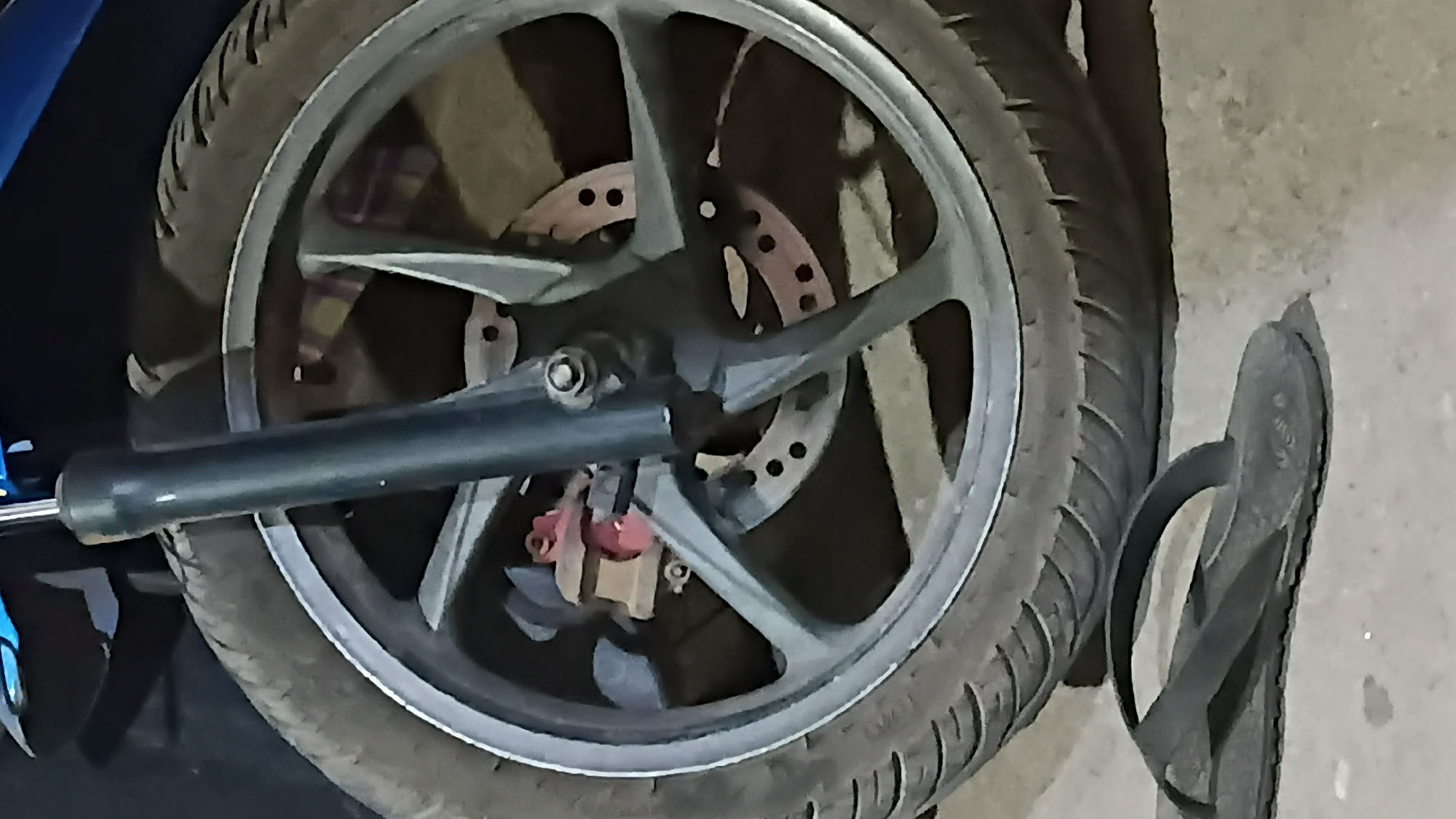 A close-up, low-angle shot captures a motorcycle wheel and a flip-flop on a concrete surface. The motorcycle's front wheel features a silver rim, a black tire with visible tread, and a grey brake disc with red brake calipers. A dark grey shock absorber is positioned vertically in the center of the wheel. The flip-flop, dark in color, lies on its side in the foreground, with its sole facing the viewer and its strap arching upwards. The background is dark and indistinct, suggesting an outdoor environment, possibly in the city of Prabumulih, Indonesia.