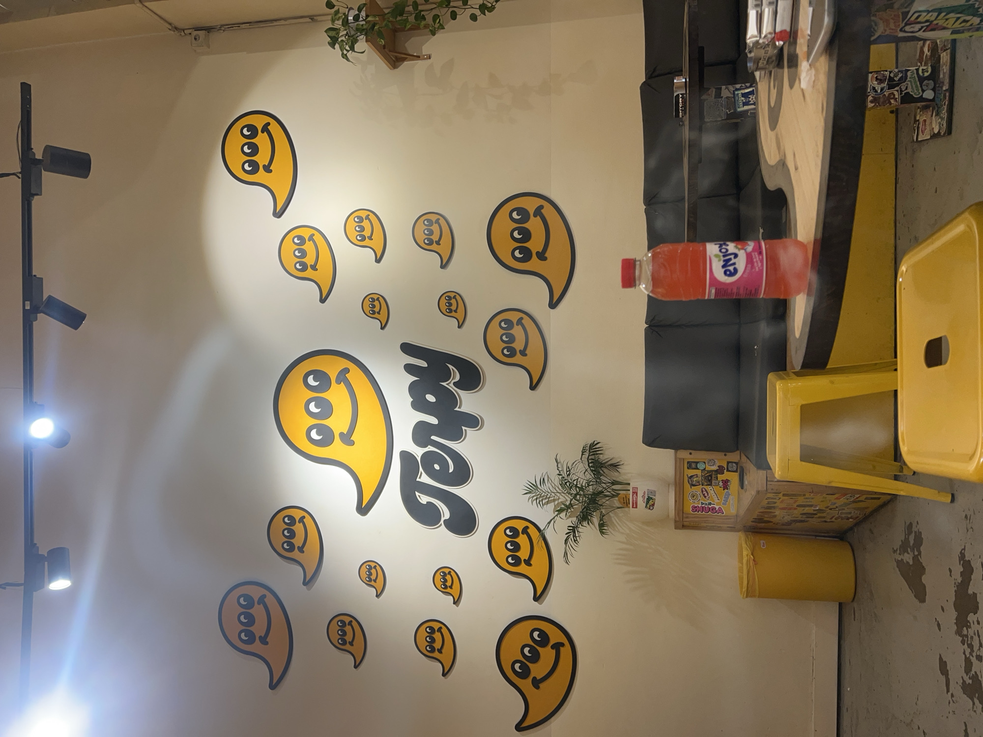 Here is a detailed description of the image:
**Environment:**
The photo appears to be taken inside a brightly lit commercial space, likely a cafe or a dessert shop.
**Objects:**
* **Wall Decorations:** The wall is the focal point, featuring a playful design with yellow, paisley-shaped figures with three eyes and smiley faces. The word "Terpy" in black, stylized lettering is prominently displayed amidst the figures.
* **Furniture:** A dark-colored cushioned seating area is visible along the right side of the frame. A yellow stool or chair is placed near a wooden table with stickers.
* **Lighting:** A tall, black lighting fixture with multiple adjustable spotlights is on the left side of the frame.
* **Miscellaneous Items:** A bottle of "enjoy" juice, a plant, and a trash can are also present.
**Details:**
* The walls are painted in a light color, possibly white or cream, providing a bright and clean backdrop for the decorations.
* The floor appears to be a textured surface, possibly concrete.
* The overall style of the space seems modern and playful.
**Barcelona, Spain:**
Without additional information (e.g., signage, distinctive architectural elements, or local products), it is impossible to conclusively determine if the location is in Barcelona, Spain. However, the modern and design-conscious aesthetic could be aligned with trends found in contemporary spaces in Barcelona.