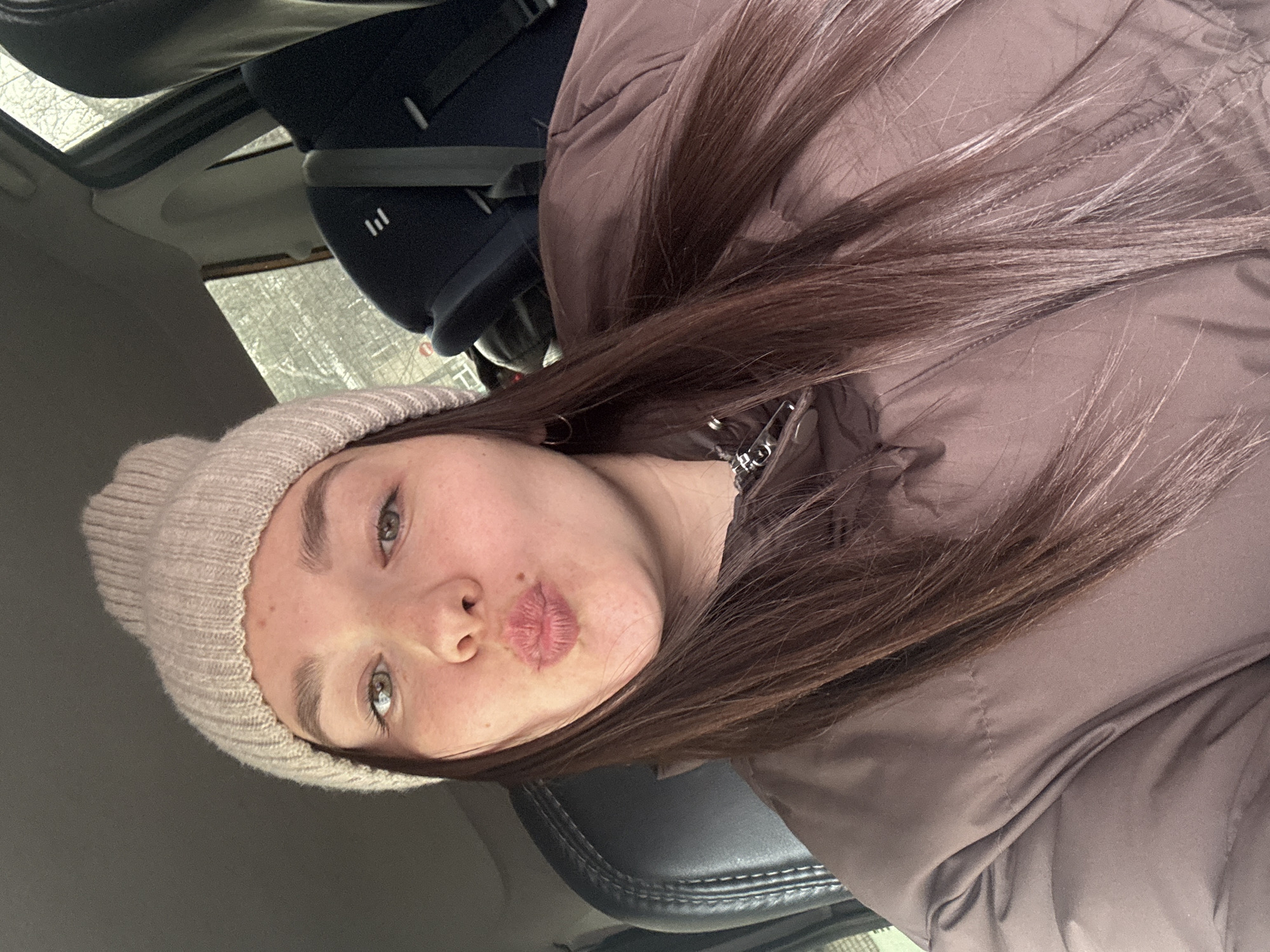 A young woman is captured in a close-up selfie, oriented horizontally as if taken inside a car. She has long, straight, dark brown hair that falls over her shoulders. Her light green or hazel eyes are open, and her lips are playfully pursed in a kissing gesture, conveying a cheerful and casual mood. She is wearing a light beige knitted beanie hat and a dark brown or taupe puffer jacket, suggesting cold weather. A silver-colored chain or necklace is subtly visible near her neck.
The setting is inside a vehicle, with dark car seats visible behind her. A dark blue child car seat with a distinctive white "|||" logo is identifiable in the upper background. Through a window or sunroof, an indistinct, textured surface, possibly a frosted window or an outdoor scene, is visible. The scene appears to be taking place during the daytime in Chaikovsky, Russia, aligning with the winter clothing. The overall impression is a candid, personal moment.