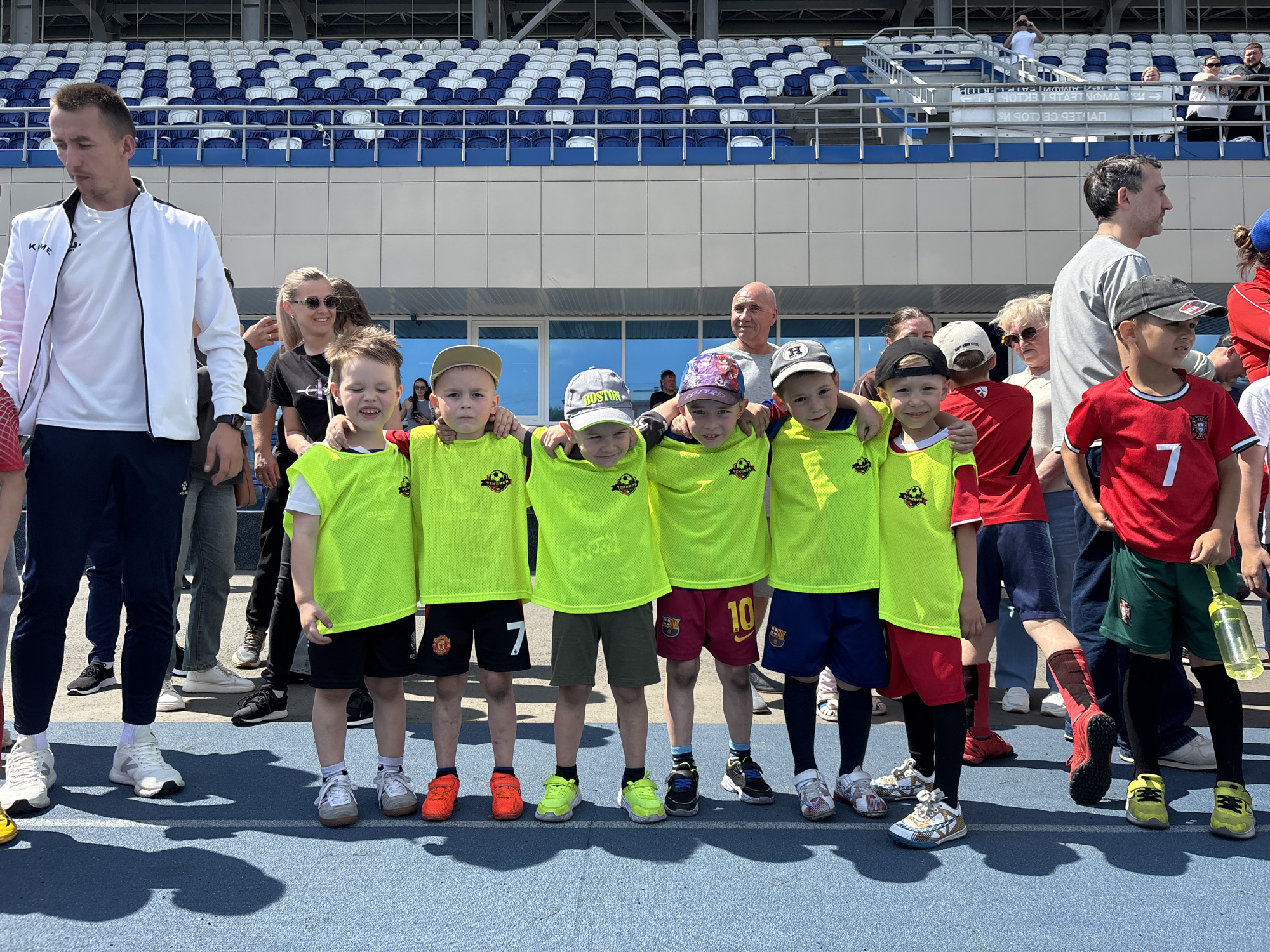 A group of young boys, some wearing bright yellow vests over soccer uniforms, pose for a photo on a blue track in front of stadium seating. A man in a white jacket and several other adults stand behind them. The stadium appears to be part of a sports complex in Izhevsk, Russia, possibly the Udmurt Republic Stadium. The day is sunny, casting shadows on the track.