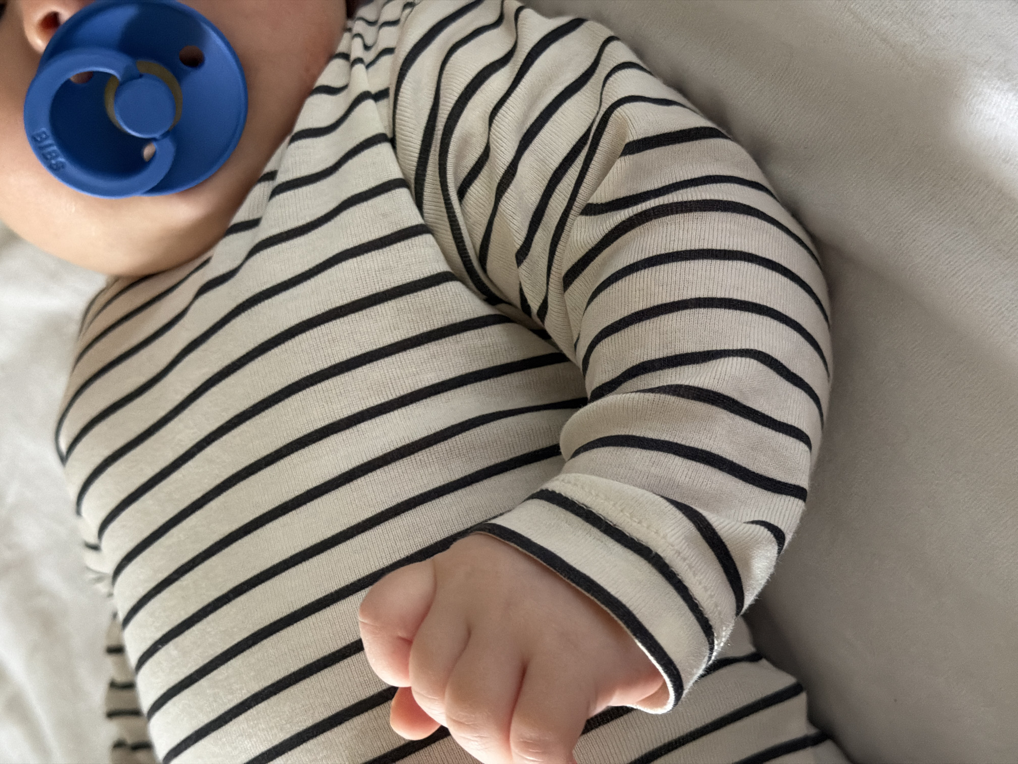 The image captures a close-up, high-angle view of a baby resting. The baby's head, partially visible on the left, features a prominent blue pacifier. The pacifier, a BIBS brand, has "BIBS" embossed on its outer shield. The baby is wearing an off-white or cream-colored long-sleeved garment with horizontal black stripes. The right arm and hand of the baby are visible, with the small hand gently curled. The baby appears to be lying on a light-colored, possibly white or cream-colored, soft fabric surface, suggesting a bed or sofa. The overall setting is indoors, with soft, diffused lighting. The activity depicted is the baby resting or sleeping peacefully. No specific time of day or outdoor weather can be determined from the image.
