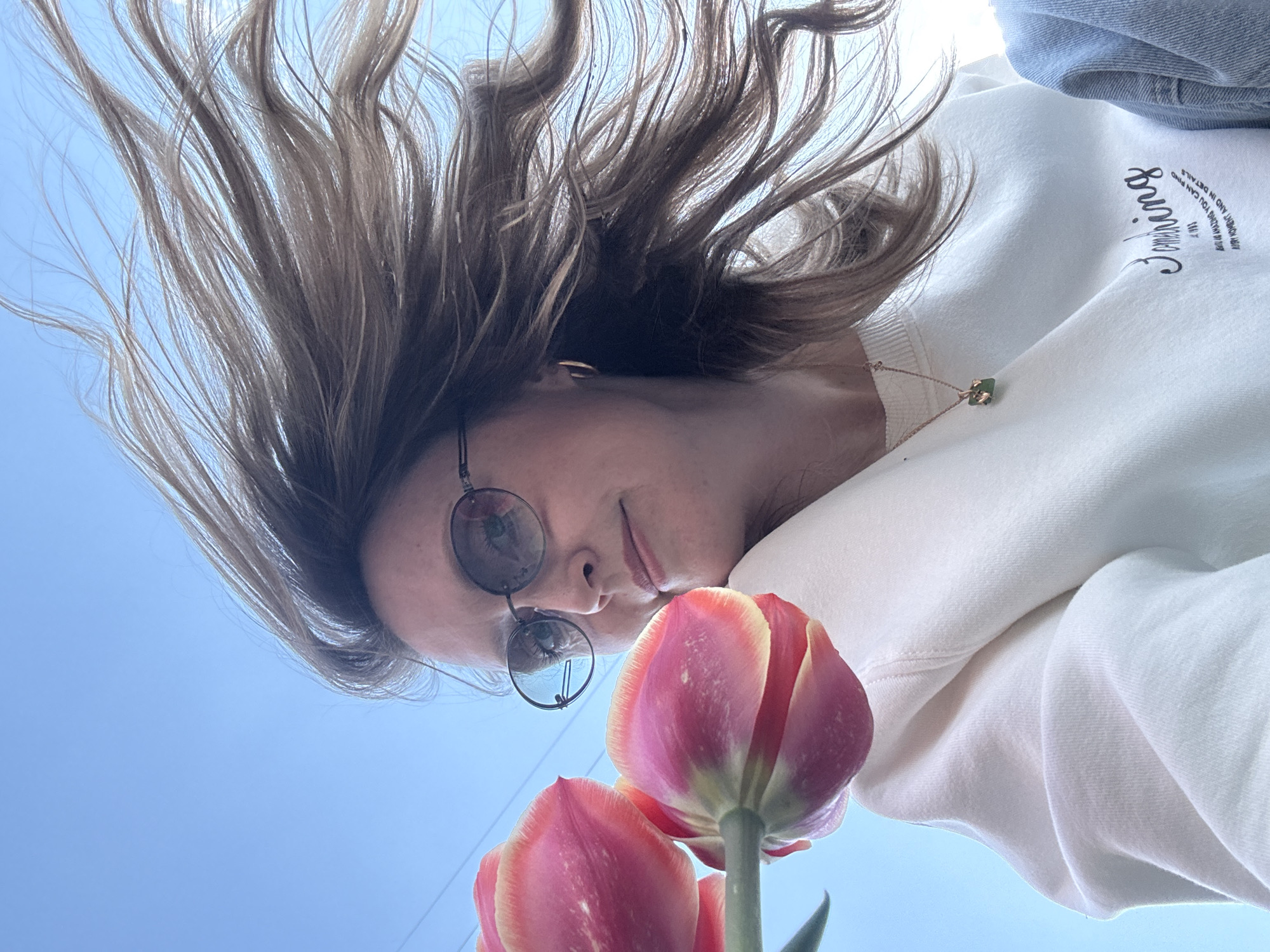 The image is taken from a low angle, looking up at a woman wearing round glasses and a white sweatshirt with script on the chest. The woman's hair is brown and blowing in the wind. In the foreground, there are two red and yellow tulips. The background is a bright blue sky. The location is unknown and cannot be determined from the image. There is no visible evidence that the photograph was taken in Chaikovsky, Russia.
