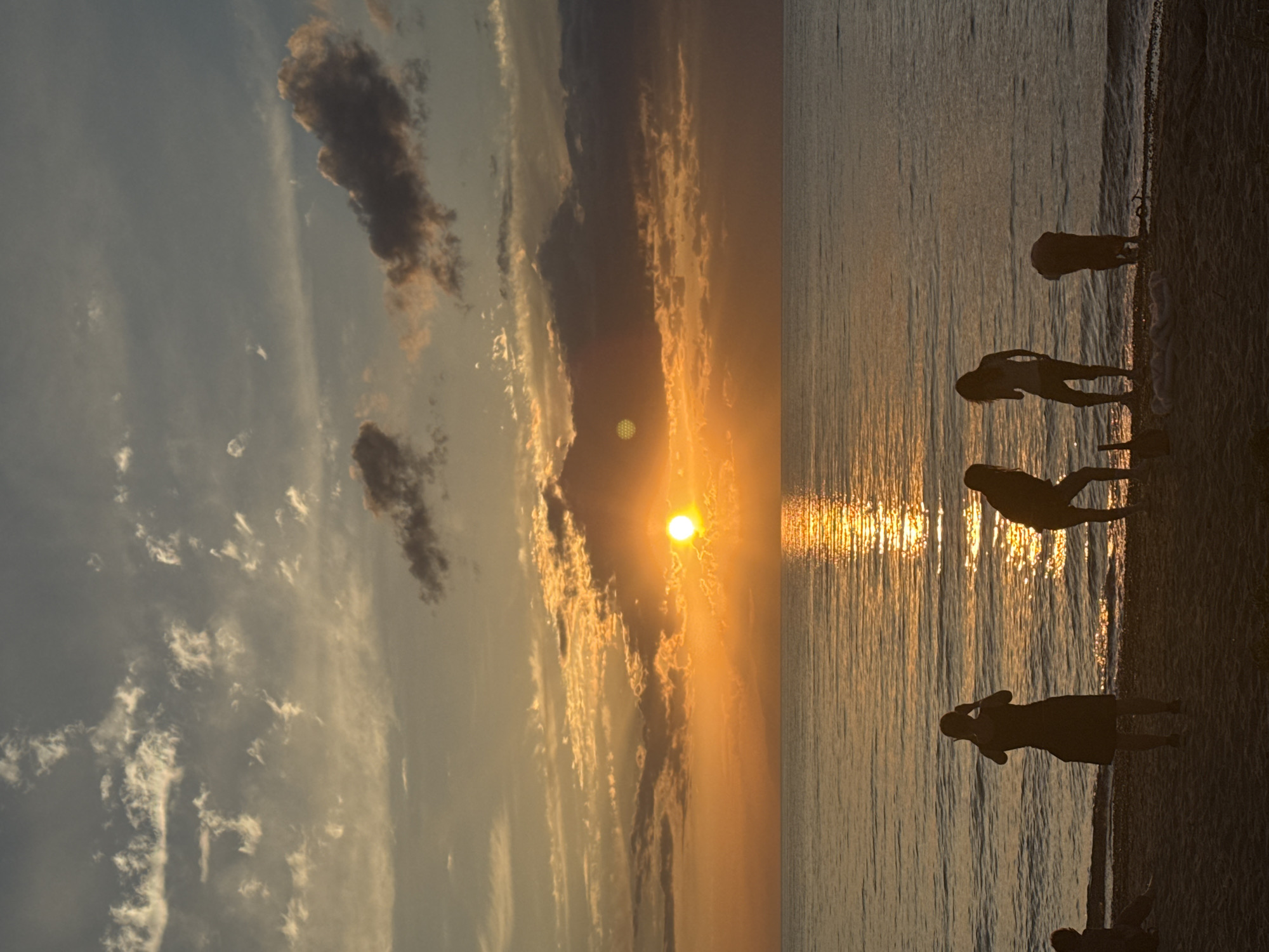 The image depicts a serene sunset over the body of water in Beshbk, Kyrgyzstan. The sky is a canvas of vibrant orange and yellow hues, with scattered clouds adding texture and depth. The sun, a bright orb, casts a shimmering reflection on the calm surface of the water. On the sandy shore in the foreground, several individuals are silhouetted against the glowing sky. One person stands with their back to the camera, appearing to take a photograph. Another person is bent over, possibly engaging in an activity on the beach. A third person stands looking out at the water, while a fourth is seated further down the shore. The overall scene evokes a sense of tranquility and natural beauty, characteristic of a peaceful evening by the water.