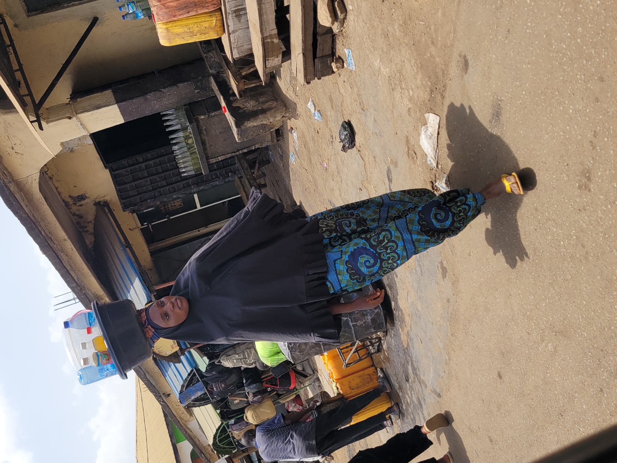 A woman in Jos, Nigeria, wearing a dark hijab and a blue and green patterned dress, is walking on a concrete surface. She is carrying a black basin on her head containing water bottles and a yellow container. The background includes a building with various items stacked, and a man in a striped shirt is also present. The location appears to be a street with various objects and debris scattered on the ground.