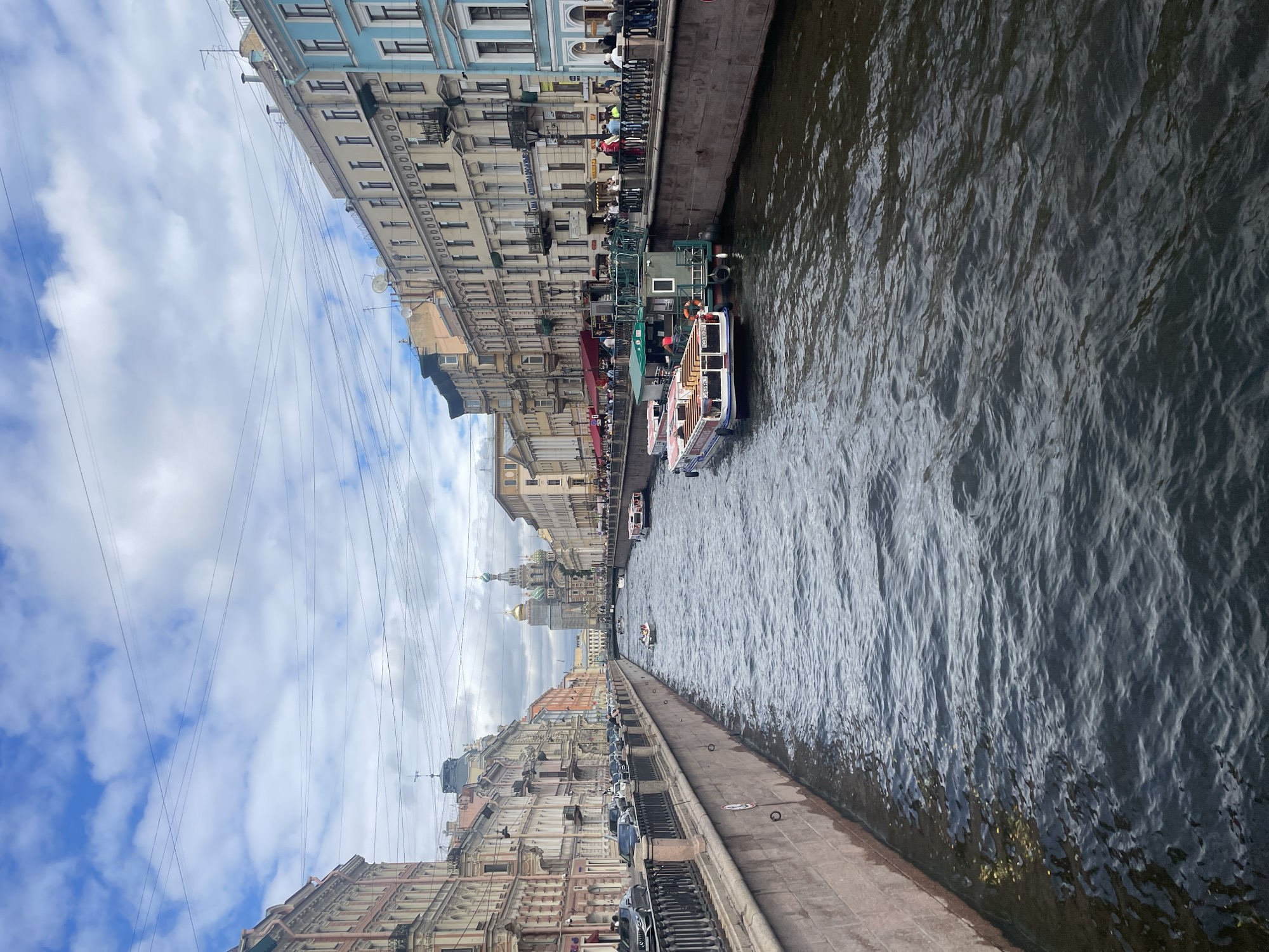 This is a vertical, eye-level, outdoor, medium shot of a canal in Saint Petersburg, Russia, likely on a cloudy day with patches of blue sky. The canal, lined with stone embankments, is filled with dark, rippling water. Several tour boats are visible on the canal, some docked and others in motion. The buildings on both sides of the canal are multi-story, with a variety of architectural styles, featuring ornate facades and numerous windows. Some of the buildings are painted in muted tones, while others are brightly colored, such as a prominent light blue building on the right. A pedestrian walkway runs along the canal bank on the right side, with people strolling and standing. A row of cars is parked on the left side of the canal, separated from the water by a black metal railing. In the distance, at the end of the canal, the golden domes of a large church, identified as the Church of the Savior on Spilled Blood, are visible, serving as a landmark. Overhead, a complex network of electrical wires crisscrosses the sky.