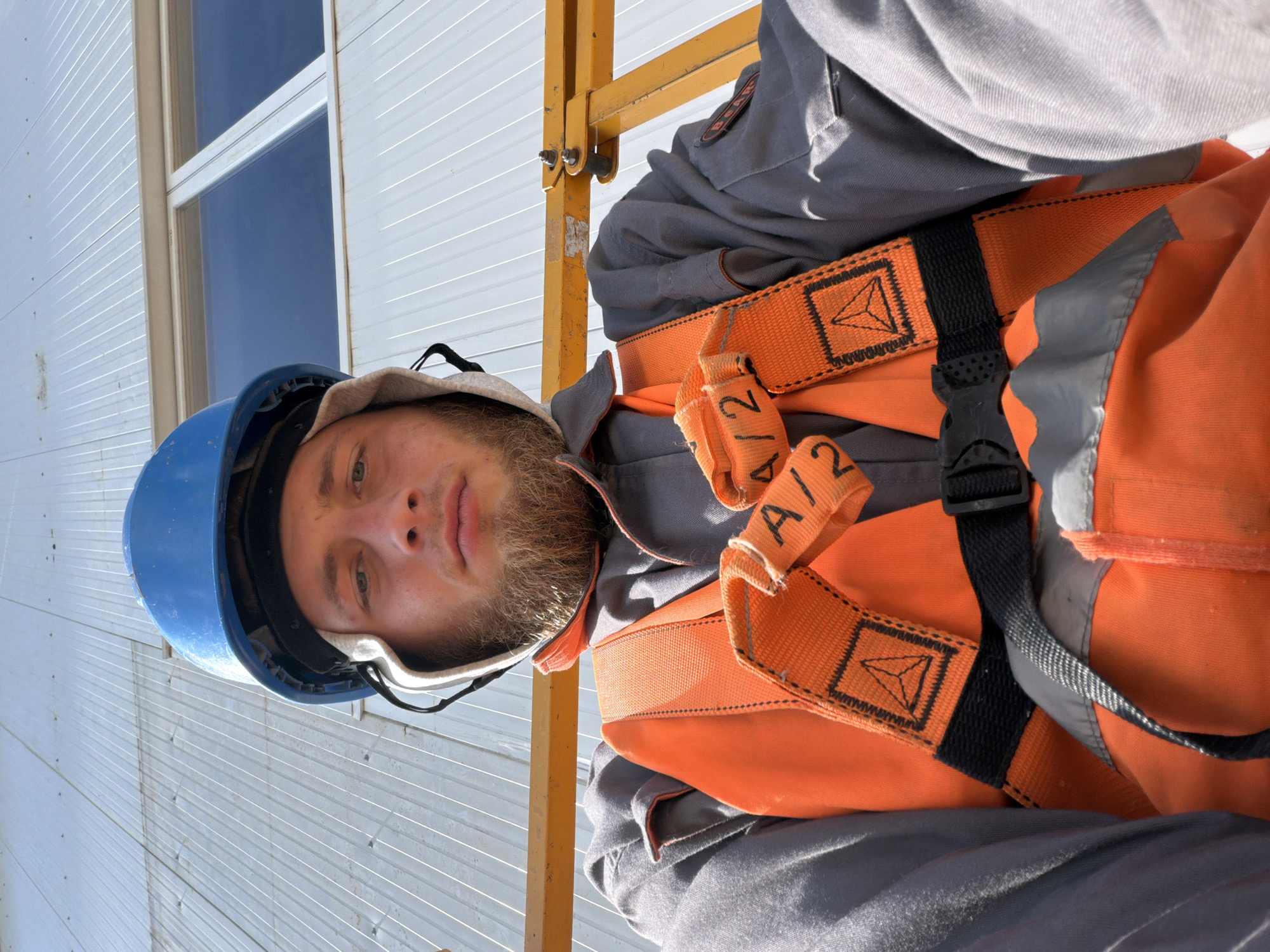 The image shows a construction worker wearing a blue hard hat, a grey uniform, and an orange safety vest with a harness. The worker has a beard and is looking at the camera. The background is a white building with horizontal lines and windows. A yellow metal beam is visible in the foreground. The image is taken from a low angle, looking up at the worker and the building. The scene suggests the worker is at a construction site. The city of Wilkowisko, Poland is mentioned, implying the location of this construction site.