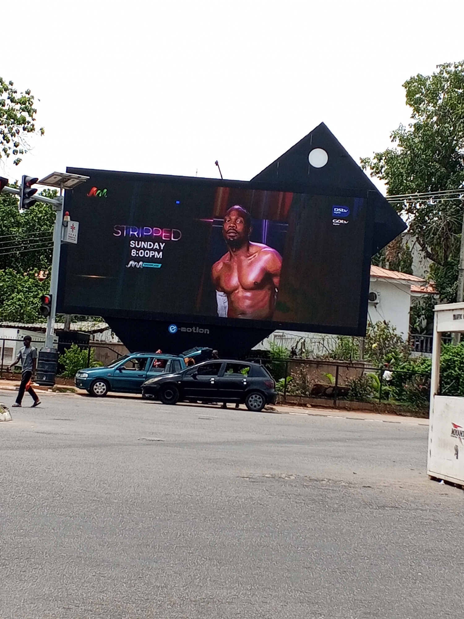 The image shows a large, digitally illuminated billboard displaying an advertisement for a program called "Stripped" on the M-Net Showcase channel, airing Sundays at 8:00 PM. The billboard features a muscular man without a shirt. Above the billboard is a small triangular architectural detail, possibly part of the billboard's support structure, with a circular opening.
Below the billboard, there's a teal-colored compact car and a black hatchback parked near a street. A man wearing a grey shirt and dark pants is walking along the sidewalk near the street. Traffic lights are visible on the left side of the image.
The environment includes trees and other greenery in the background. A white building is partially visible behind a fence and vegetation to the right of the billboard. A white advertisement sign is visible on the right side of the frame.
The location is identified as Abuja, Nigeria, based on the billboard's content and other visual cues. The advertisement is for a program on an African television channel.