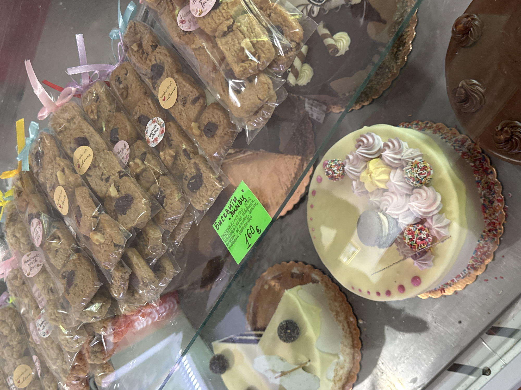 The image shows a display case filled with baked goods, likely in a bakery. In the foreground, there are several cakes and pastries. One cake is light yellow with a light blue macaron and pink meringue swirls on top, decorated with sprinkles around the base. To the left of this cake is a slice of a white-frosted cake with a blackberry on top. Another cake, visible to the right, has dark chocolate frosting and is decorated with piped cream swirls and chocolate wafer rolls. In the background, rows of individually packaged cookies, tied with ribbons, are stacked. Some of these cookies have small round labels that read "handmade with love." A bright green price tag is also visible on a glass shelf, displaying Cyrillic text and the price "160". The overall environment suggests a commercial food establishment. This display is located at the "Bakery and Confectionery" in Zheravna, Bulgaria.