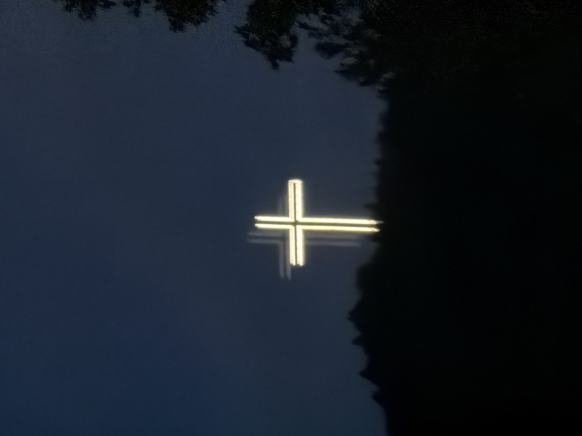 A nocturnal shot shows a brightly lit cross reflected on water. Some dark shapes, possibly foliage or trees, are visible at the top of the image. The right edge of the frame is obscured by a very dark, indistinct mass. It is not possible to determine the location within Blagoevgrad or to provide specifics about people, actions, or interactions.