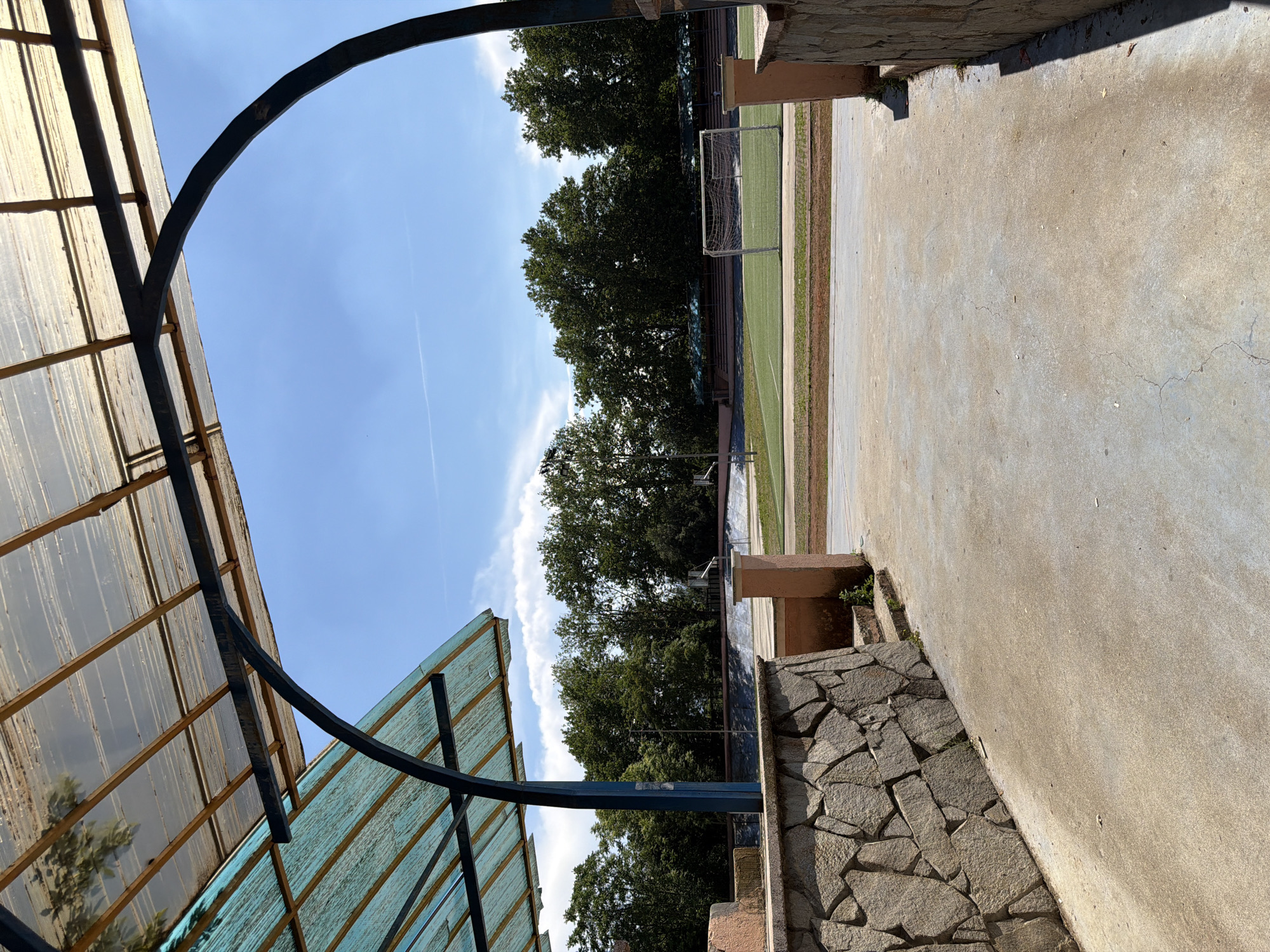 The image captures an outdoor view of a sports facility, likely a stadium or sports complex, located in Pazardzhik, Bulgaria. In the foreground, a concrete walkway leads towards an open area. To the left and right, stone walls are visible, partially obscured by a structure with a transparent, corrugated roof supported by metal beams. Beyond this structure, a green field with a soccer goal is prominent. Trees form a dense line behind the field, and a clear blue sky with scattered clouds stretches above. The overall scene depicts a sunny day at a sports venue.