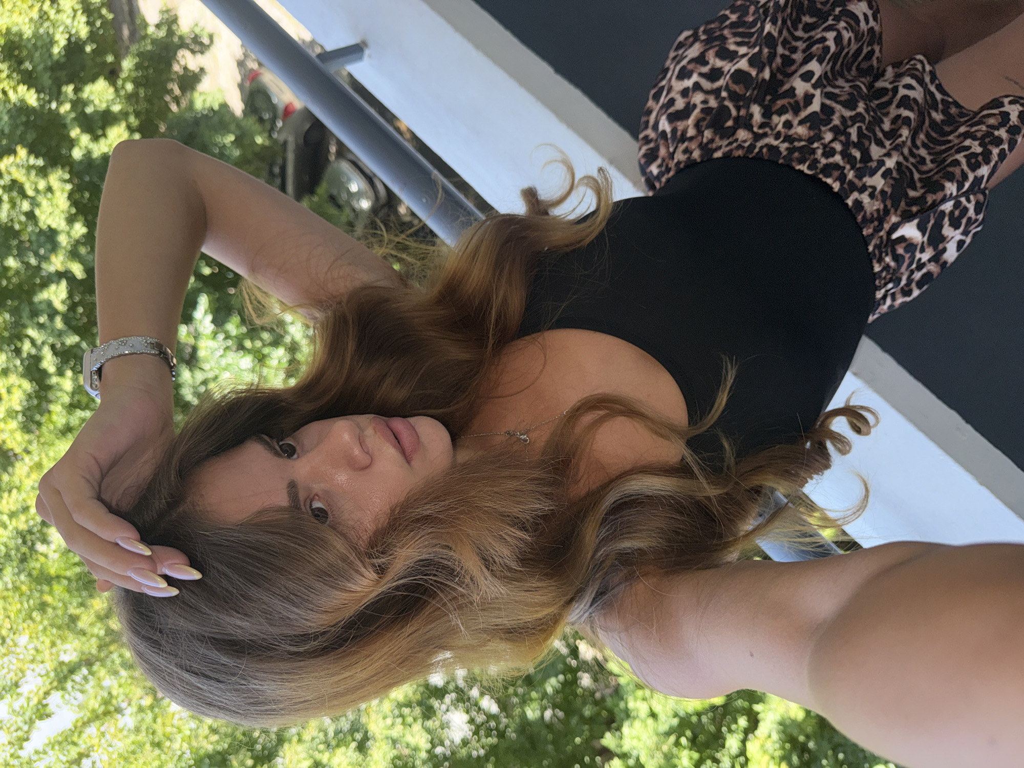 This media file is a portrait of a young woman lying on her side, with her head propped on her hand and her gaze directed towards the camera. She has long, wavy, light brown hair that falls around her face and shoulders. Her skin appears to be lightly tanned, and she is wearing a black tank top and a leopard-print skirt. A silver watch with a white band is visible on her left wrist. A delicate silver necklace is also visible around her neck.
The background suggests an outdoor setting, with lush green foliage, possibly trees, creating a soft, blurred backdrop. A white railing and a dark gray surface are also present, hinting at a balcony or patio. The lighting suggests it is daytime, likely sunny, given the bright highlights on her skin and hair and the strong contrast in the background. The overall mood is relaxed and casual. The image was taken in Plovdiv, Bulgaria, and the foliage and railing might be indicative of the architectural style common in the region. There is no visible text in the media file.