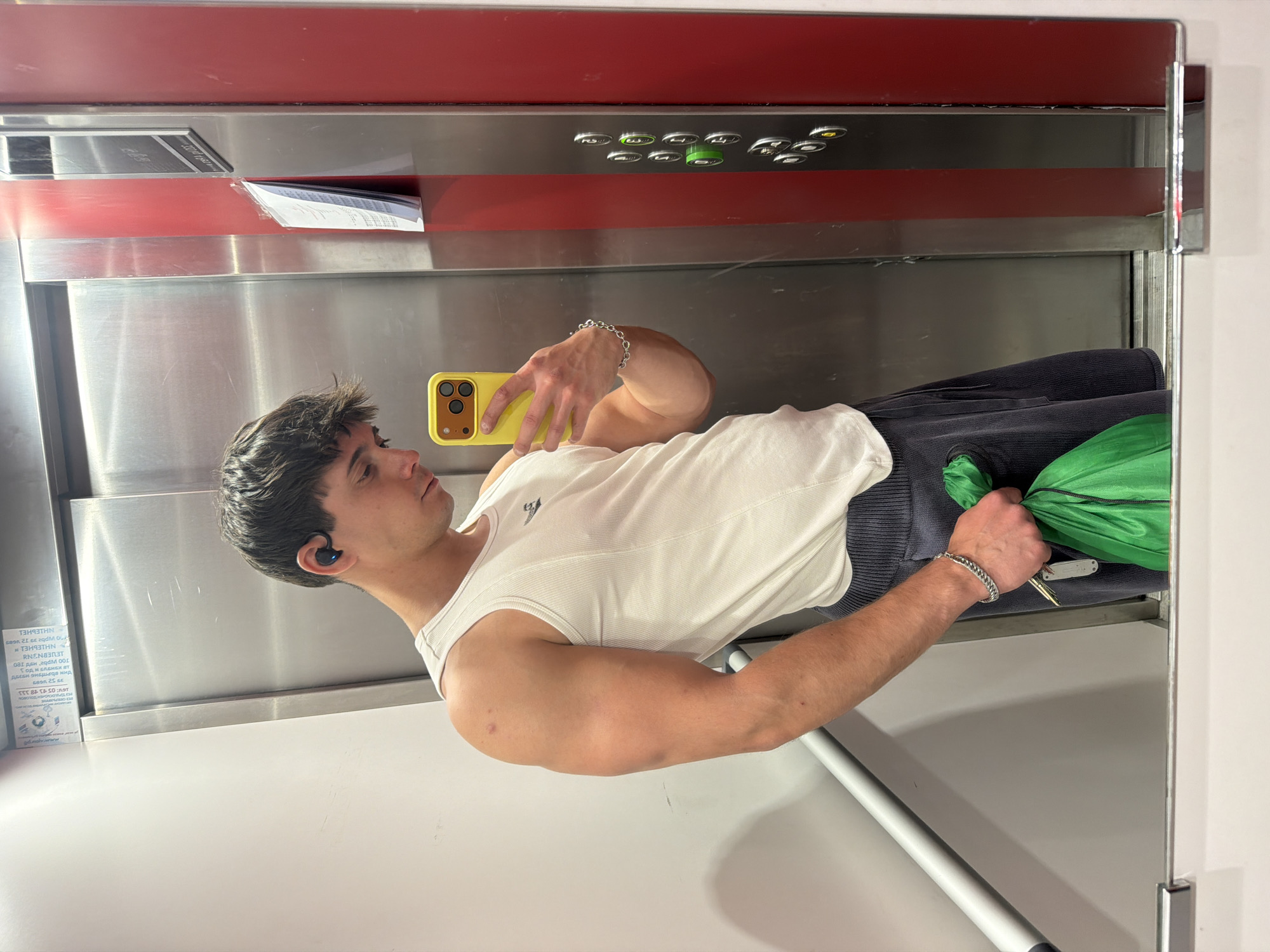 A young man is captured in a mirror selfie, with the image oriented 90 degrees clockwise, causing him to appear horizontally in the frame. He is looking at his yellow-cased smartphone, which he holds in his right hand to take the picture. He wears a white ribbed tank top, dark bottoms, and a black wireless earbud in his left ear. A silver chain bracelet adorns his right wrist, while his left hand holds a set of keys and a bright green bag or garment.
The setting appears to be an elevator or a changing room in Sofia, Bulgaria, characterized by polished metallic walls and a prominent red horizontal strip running across the upper portion of the background. Above this red strip, a control panel with several silver and green circular buttons is visible. On a lower metallic surface to the left, a white label with partially obscured and inverted text is present.
The visible text, reflected and partially legible from the label, includes: "16:05 43 48 3", "ДБЕБИ ЭНБДа нид", "ГОДИ БЛЕНЕХ ВТ", "LED", "REN", "TOO Wpbe Hd Jeo", "NHIEUHEL", "Wpbe 29 12 vees", "OLA", and "T".