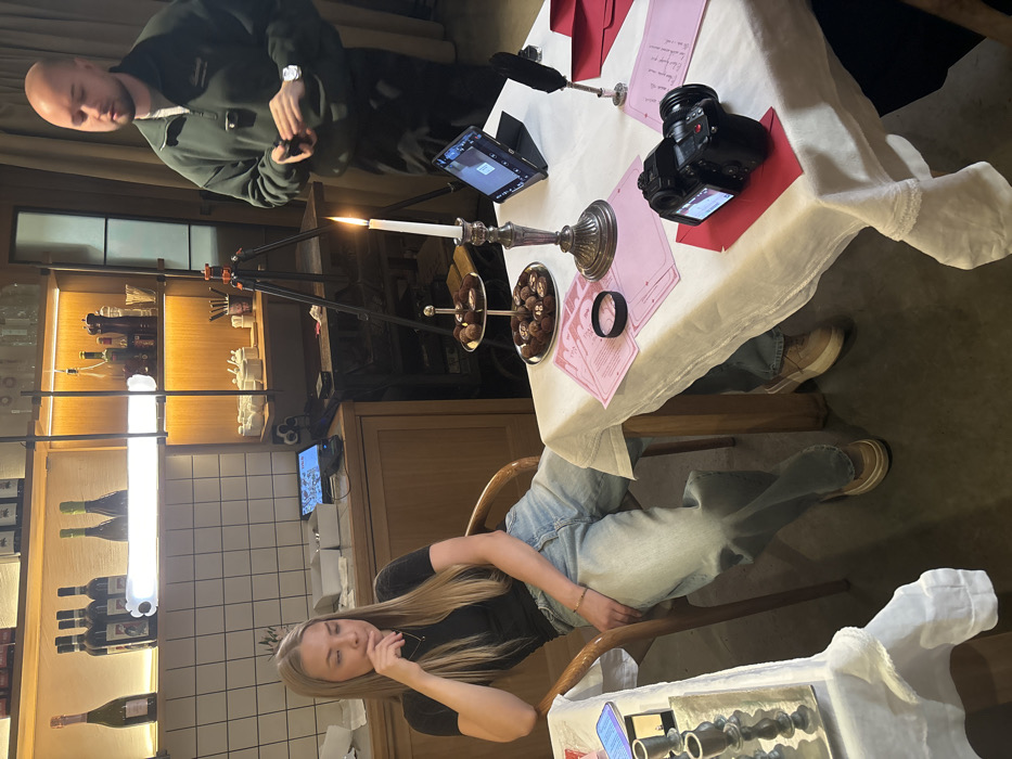 The image depicts an indoor scene in Saint Petersburg, Russia, within what appears to be a commercial establishment such as a cafe or restaurant. A blonde woman, wearing a black short-sleeved top and light blue wide-leg jeans, is seated in a wooden chair in the foreground, resting her left hand on her chin with a contemplative expression. Her right arm is positioned across her lap. To her right, a table draped with a white tablecloth features a lit white candle in a silver candelabra, a two-tiered dessert stand filled with chocolates or truffles, several pink printed documents, a black wristband, a black digital camera resting on a red envelope, a tablet displaying an image, and a black quill pen positioned near additional red envelopes. To the left of the woman, a portion of another table with a white tablecloth and a silver candelabrum is visible.
A bald man, dressed in a dark green quarter-zip sweatshirt, stands in the mid-ground to the right, behind the table. He holds a small black device in his right hand, with a watch visible on his left wrist, and a small microphone is clipped to his sweatshirt. A black camera tripod stands between the man and the table with the lit candle.
The background features a white tiled wall section above a wooden counter. Above the counter, wooden shelves display an assortment of bottles, including wine and spirits, illuminated by concealed lighting and a horizontal fluorescent tube light. A small electronic screen or tablet is present on the wooden counter.