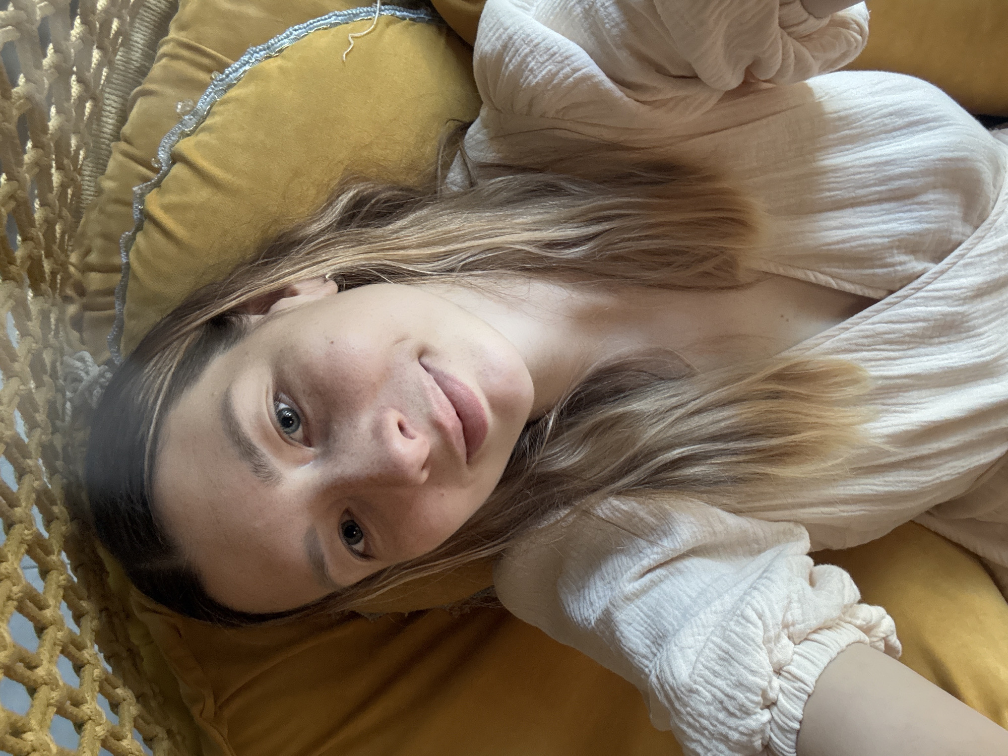 A woman with blonde hair and fair skin is lying down on a yellow cushion or pillow, wearing a white, long-sleeved, textured shirt. Part of a woven hammock or hanging chair is visible on the left side of the image. The background is blurred, but no elements indicate the location is Чёрное, Russia.