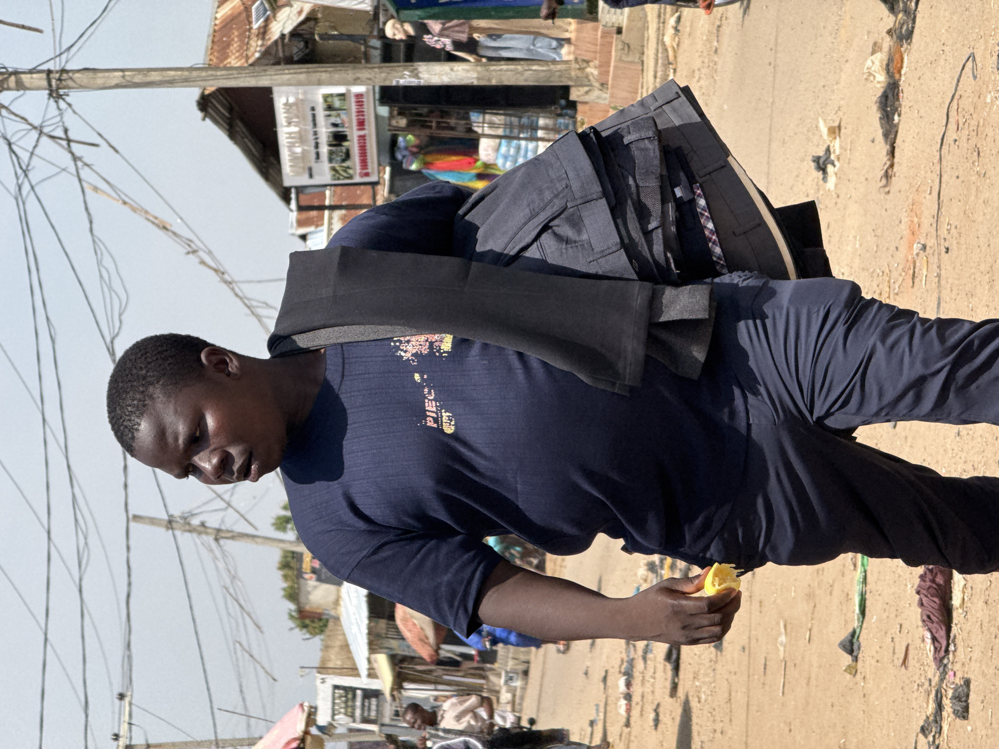 In Abuja, Nigeria, a man is seen walking in a dusty market environment. He wears a dark blue t-shirt with a graphic design on it and dark blue trousers. He is carrying a stack of dark-colored trousers or pants draped over his left shoulder, with a dark scarf or piece of cloth also on his shoulder. In his right hand, he holds a piece of yellow fruit, possibly a lemon, which he appears to be eating.
The background reveals a bustling marketplace with various stalls and buildings. A sign for "WHEEL STORE" is visible on one of the buildings, along with a phone number. Mannequins, one dressed in a pink top and blue jeans, are displayed outside a shop. The ground is covered in dirt and scattered debris, suggesting a typical outdoor market setting. Overhead, a tangle of electrical wires crisscrosses the sky, a common sight in many urban areas. The overall scene depicts a moment of daily commerce and activity in the city.