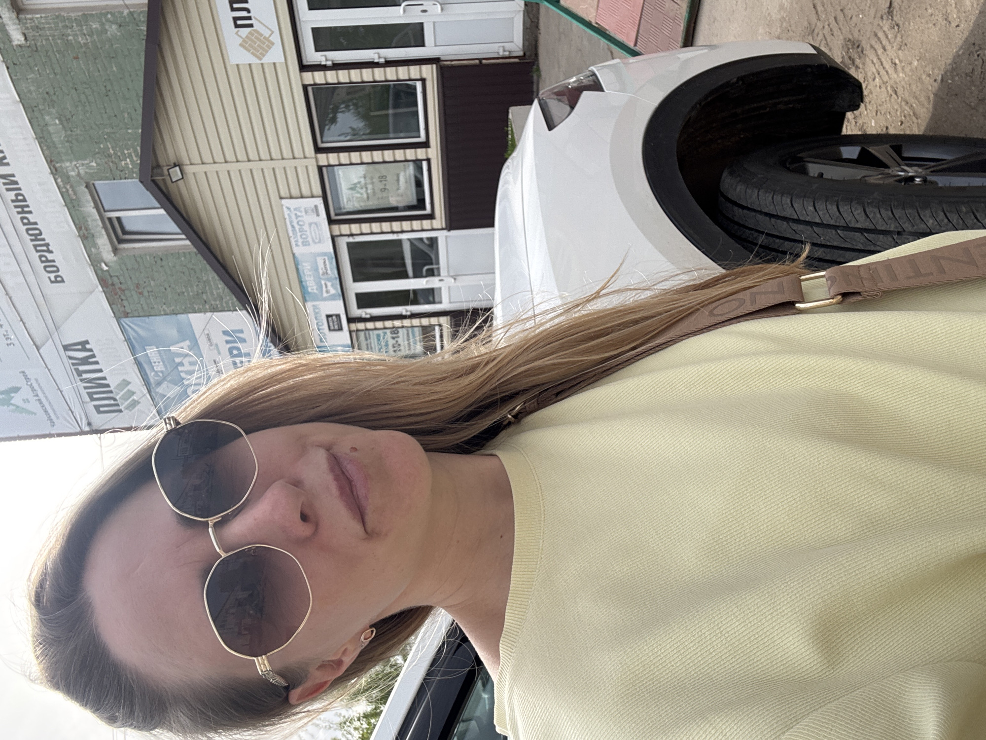 A selfie shot depicts a fair-skinned woman with shoulder-length blonde hair, wearing sunglasses and a yellow sweater, and a brown shoulder strap. Behind her and to her right is the front end of a white car. Beyond the car is a single-story building with beige siding and brown trim, featuring windows and doors. Signs with Russian text are visible on the building. Based on the signage, the location is Chaikovsky, Russia.