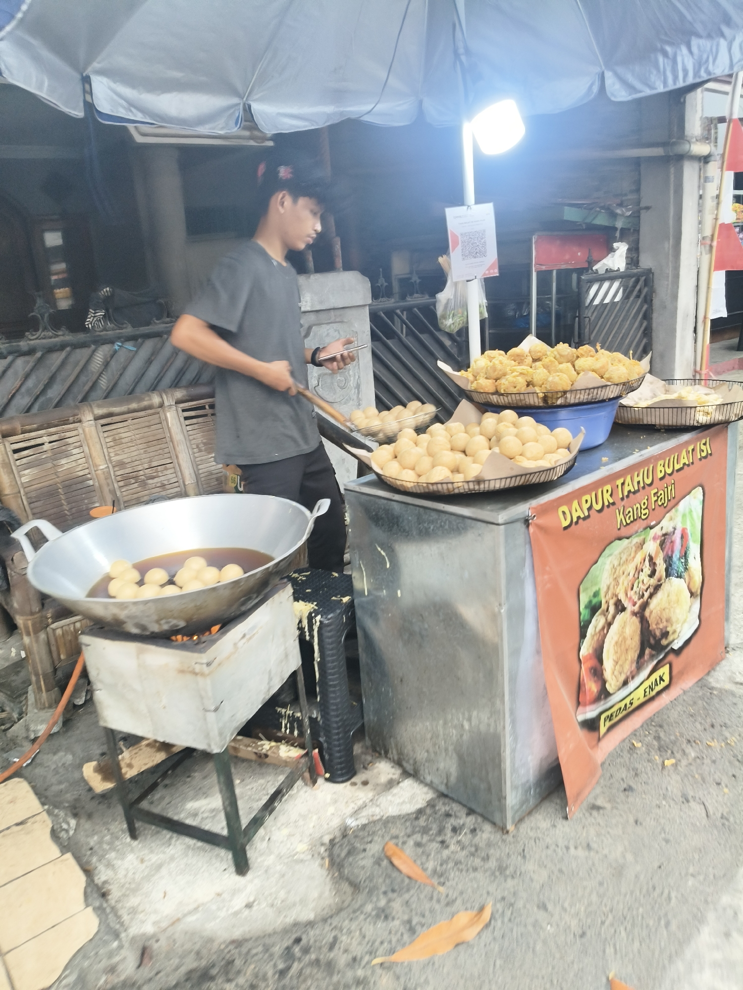 A street vendor in South Tangerang, Indonesia, is frying round, tofu-like snacks in a large wok over a charcoal stove. The vendor, a young man wearing a dark grey t-shirt and a black baseball cap, is using a wooden utensil to manage the frying process and holds a smartphone in his other hand. He is positioned under a large, white umbrella that provides shade.
In front of the vendor, a metal cart holds several baskets of fried food. Two baskets on top of the cart contain numerous golden-brown, spherical snacks. Another basket, placed to the left of the cart, appears to hold uncooked snacks. A banner on the front of the cart displays an image of fried food and text that reads "DAPUR TAHU BULAT ISI Kang Fajri PEDAS - ENAK".
The scene is outdoors, on a paved surface that appears to be a sidewalk or street. Debris, including fallen leaves, is visible on the ground. The background shows a building with a slatted wooden facade and a metal railing. A QR code is displayed on a white piece of paper, attached to a pole near the umbrella.