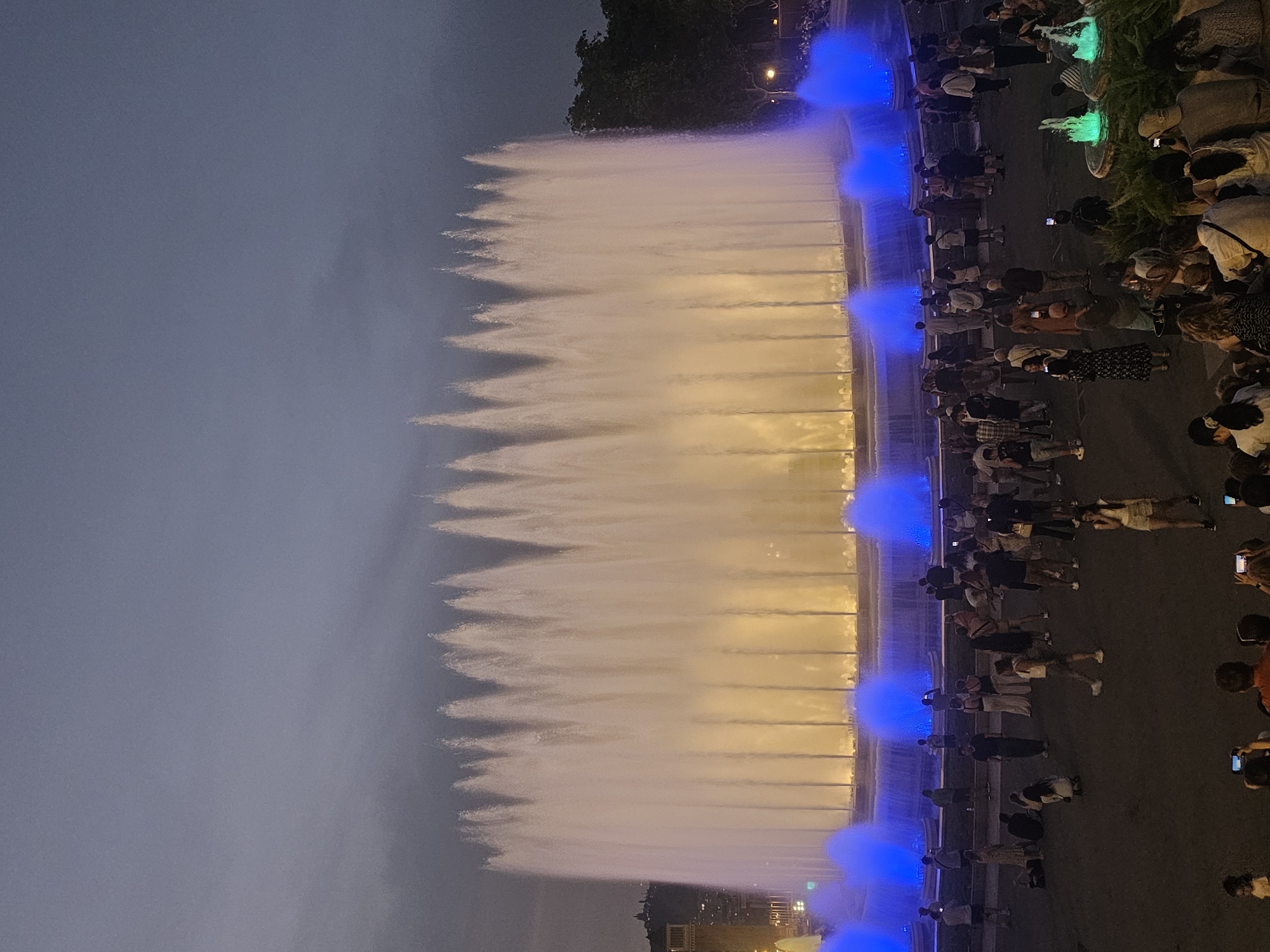 The image depicts the Magic Fountain of Montjuïc in Barcelona, Spain, at dusk. A large, illuminated fountain dominates the center, with multiple streams of water rising high into the air, lit from below with warm yellow light and accented with vibrant blue light at its base. A crowd of people, of varying ages and ethnicities, are gathered around the fountain, many of them observing the spectacle, some taking photographs with their phones. The ground is paved, and trees are visible in the background to the right. The sky is a dark, overcast blue, indicating the transition from day to night.