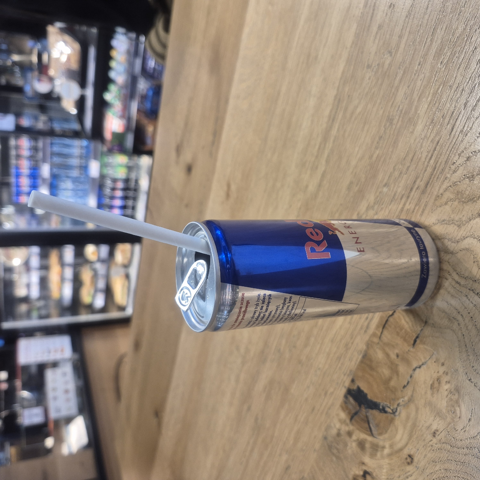 The image shows a close-up of a Red Bull energy drink can with a straw inserted. The can is primarily blue and red, with the distinctive Red Bull logo visible. The straw is white and is positioned in the opening of the can. The can is placed on a wooden surface, which appears to be a table with a visible wood grain pattern. In the blurred background, a refrigerator or display case filled with various items, likely drinks and food, can be seen. The lighting suggests an indoor setting, possibly a store or convenience store. The text on the can is partially visible and appears to be in Polish, mentioning ingredients and nutritional information like "Zawiera tauryne" (Contains taurine) and "1/46 kcal". The scene is static, focusing on the object itself. There are no people visible in the image.
